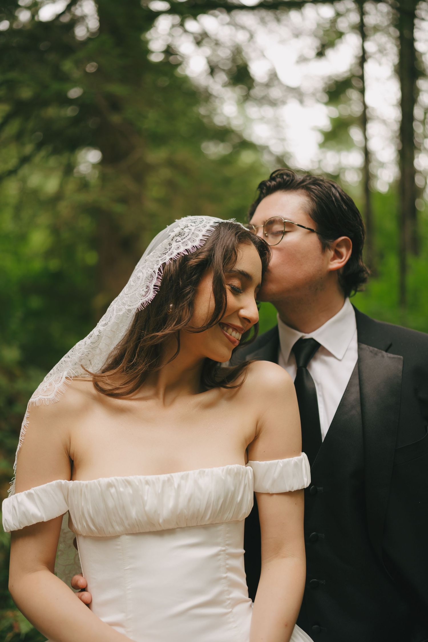 Groom in black suit kissing bride in off-shoulder white gown and lace veil, smiling in lush green forest.