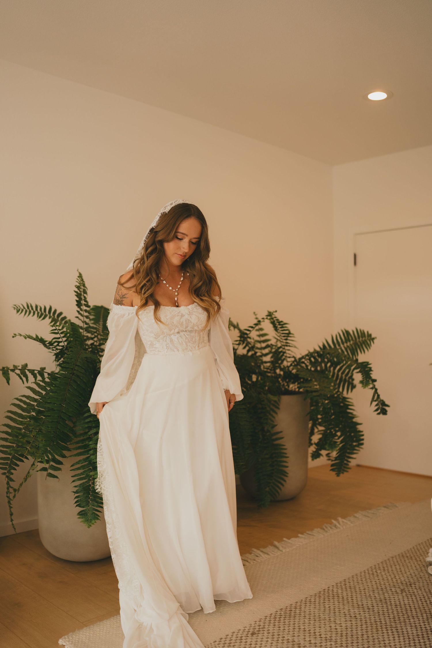 A flowing white wedding dress is displayed in a room with potted ferns and warm lighting.