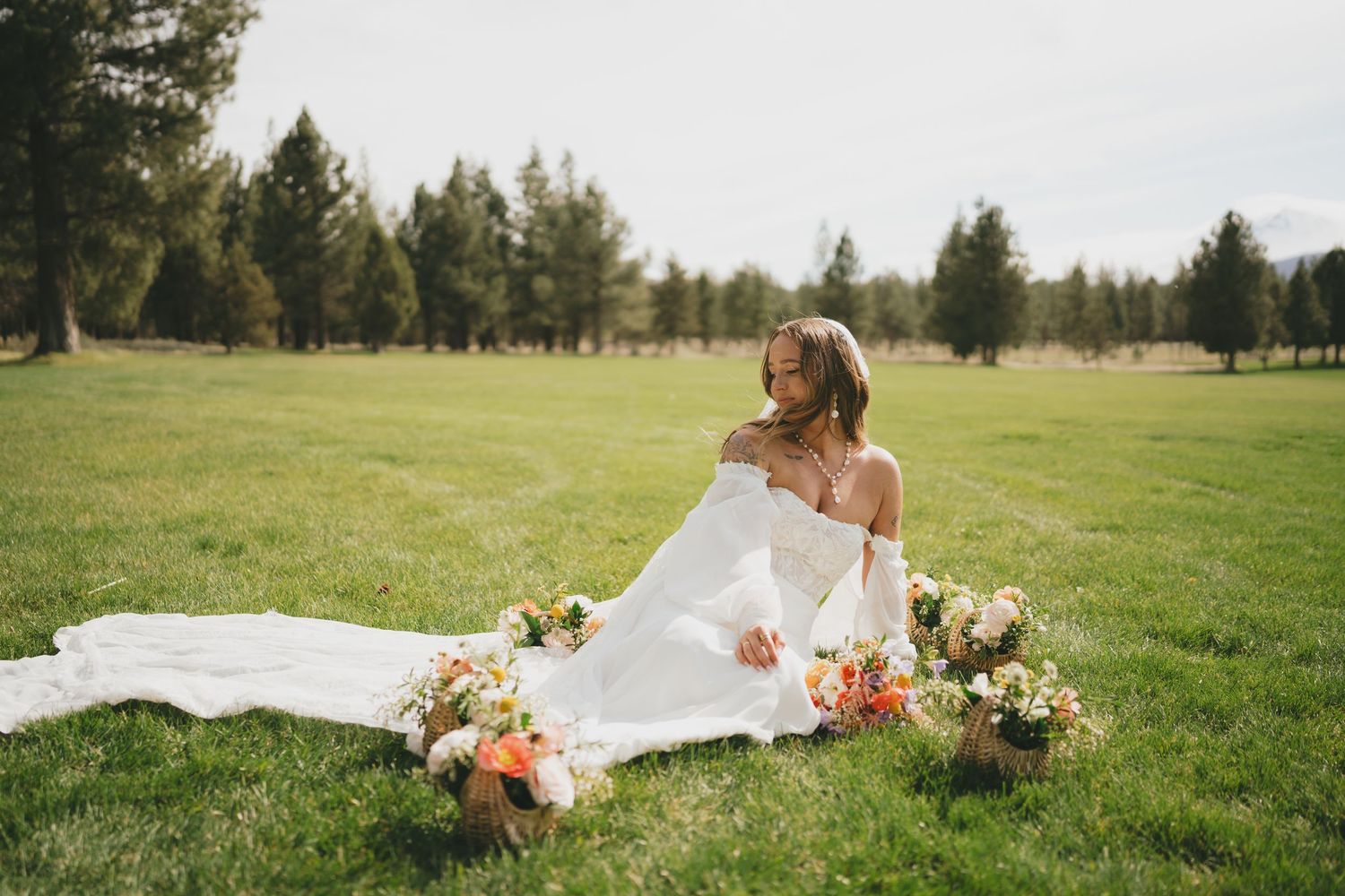 A bride in a flowing white dress sits on grass with colorful floral arrangements in an outdoor garden setting.
