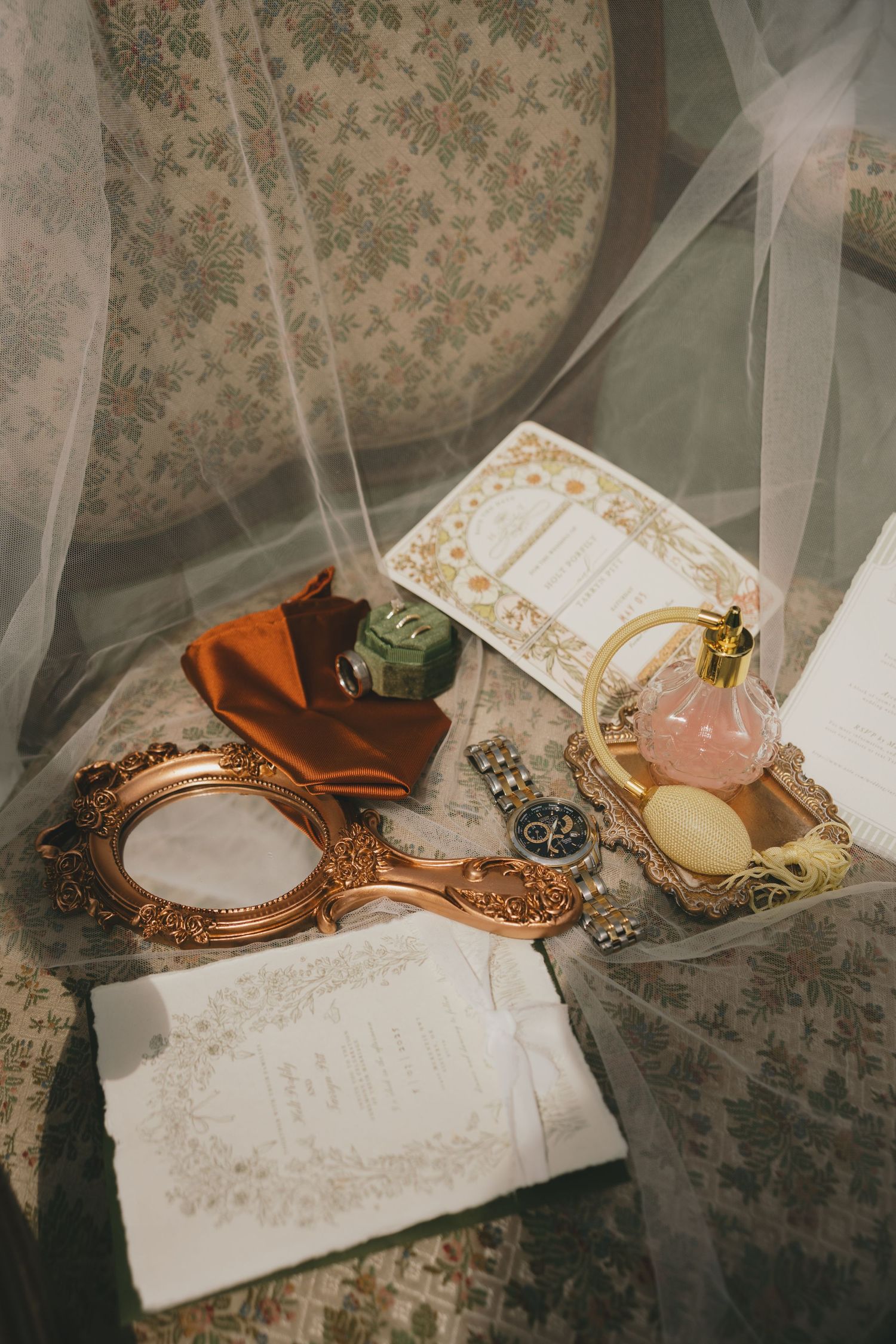 Vintage vanity setup with ornate copper mirror, perfume bottles, and floral fabric backdrop under sheer veil.