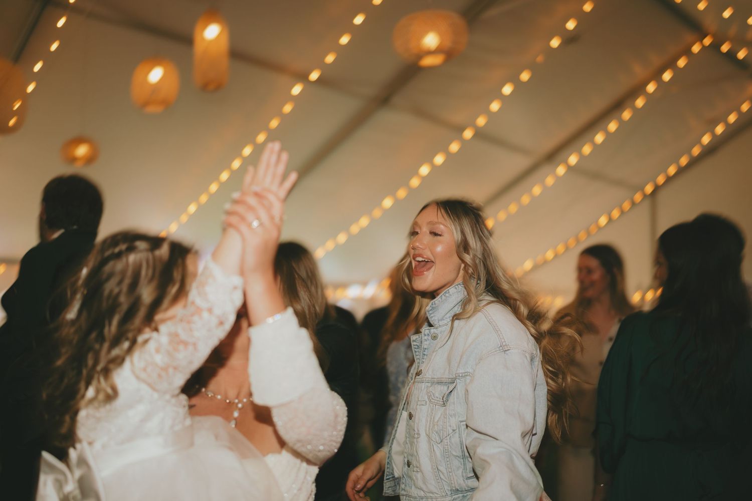 Wedding guests dance and celebrate on a dimly lit dance floor with string lights glowing overhead at a reception.