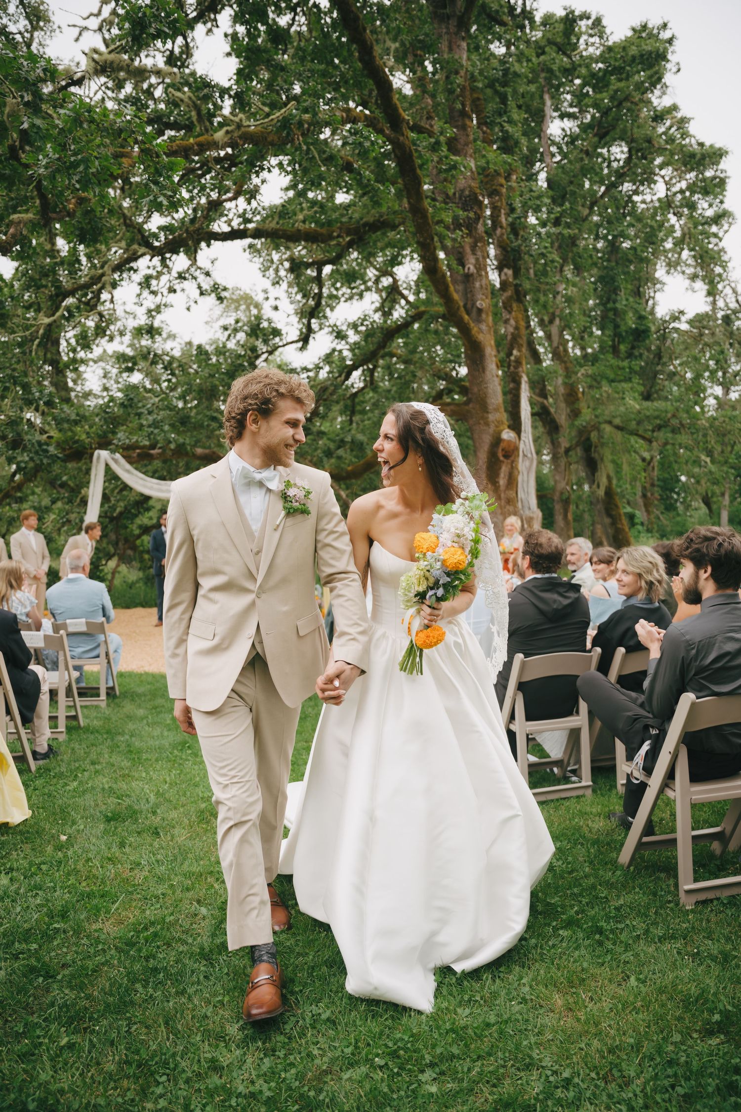 Bride and groom walk hand-in-hand down the aisle at an outdoor wedding ceremony surrounded by lush green trees.