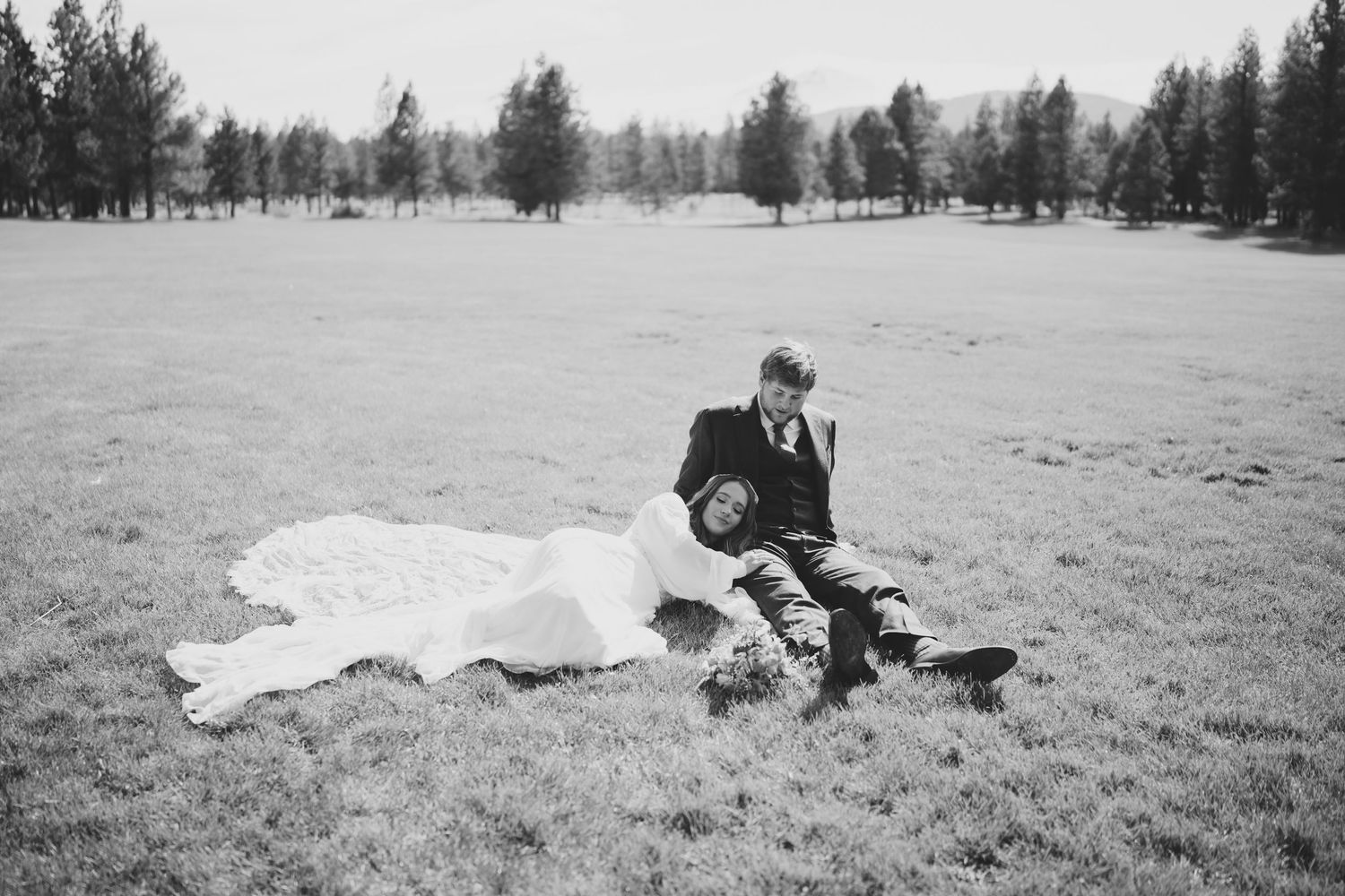 Black and white photo of a couple sitting together on a blanket in a field with trees in the background.