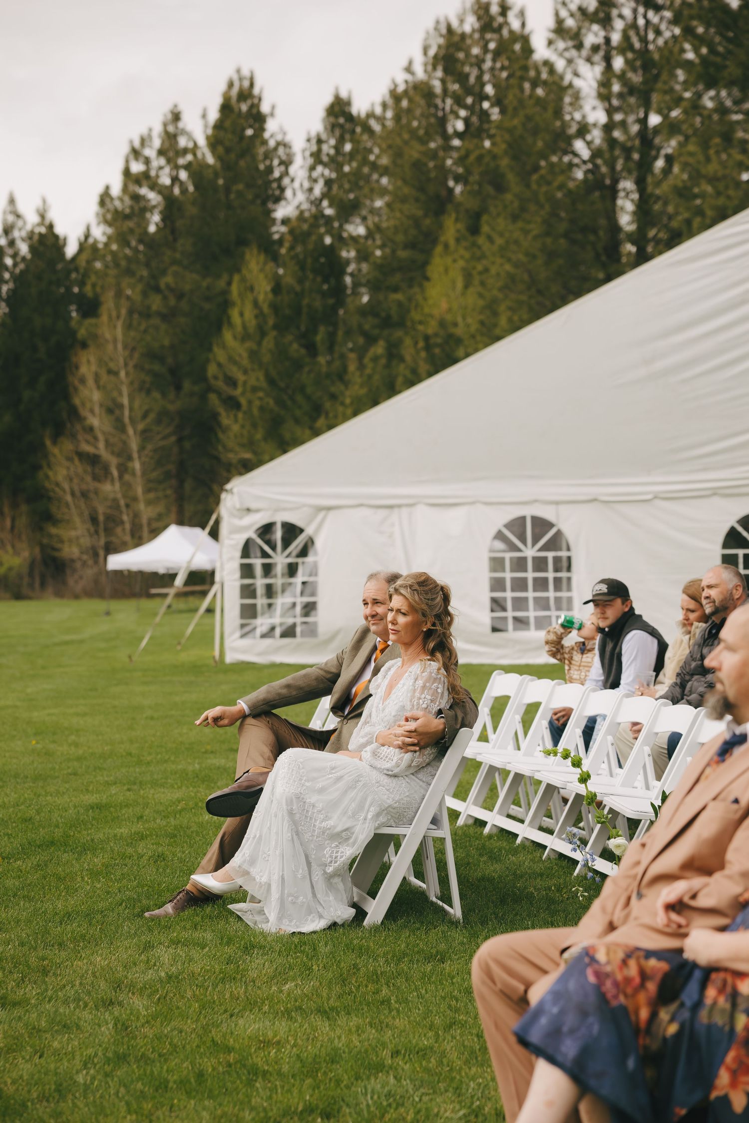 Wedding guests sit in white chairs on a green lawn with white tents and pine trees in the background.