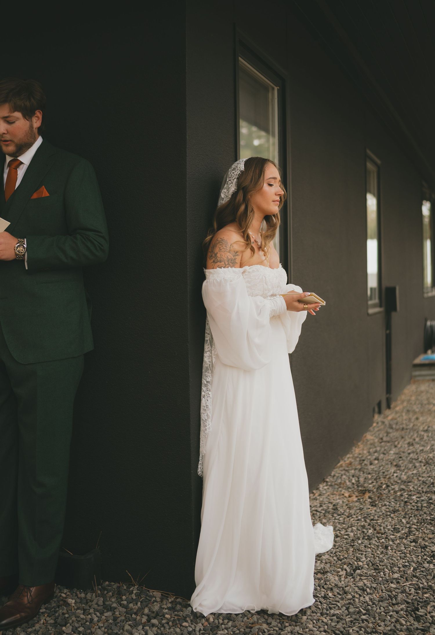 Bride in flowing off-shoulder white dress and groom in dark green suit stand separately against dark wall during first look.