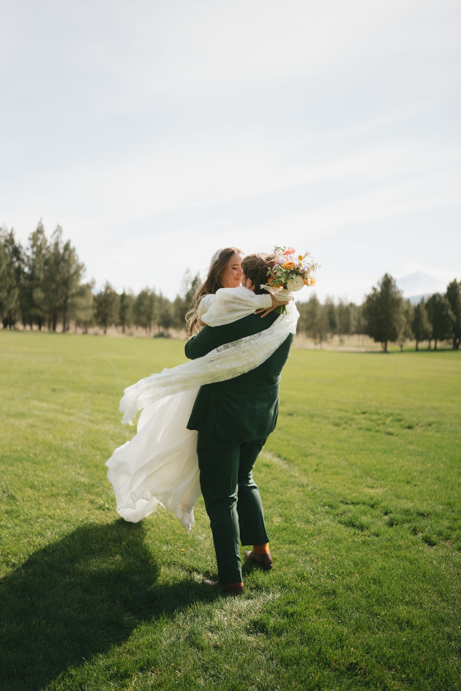 A couple embraces in a joyful spin on a grassy field with trees in the background during an outdoor celebration.