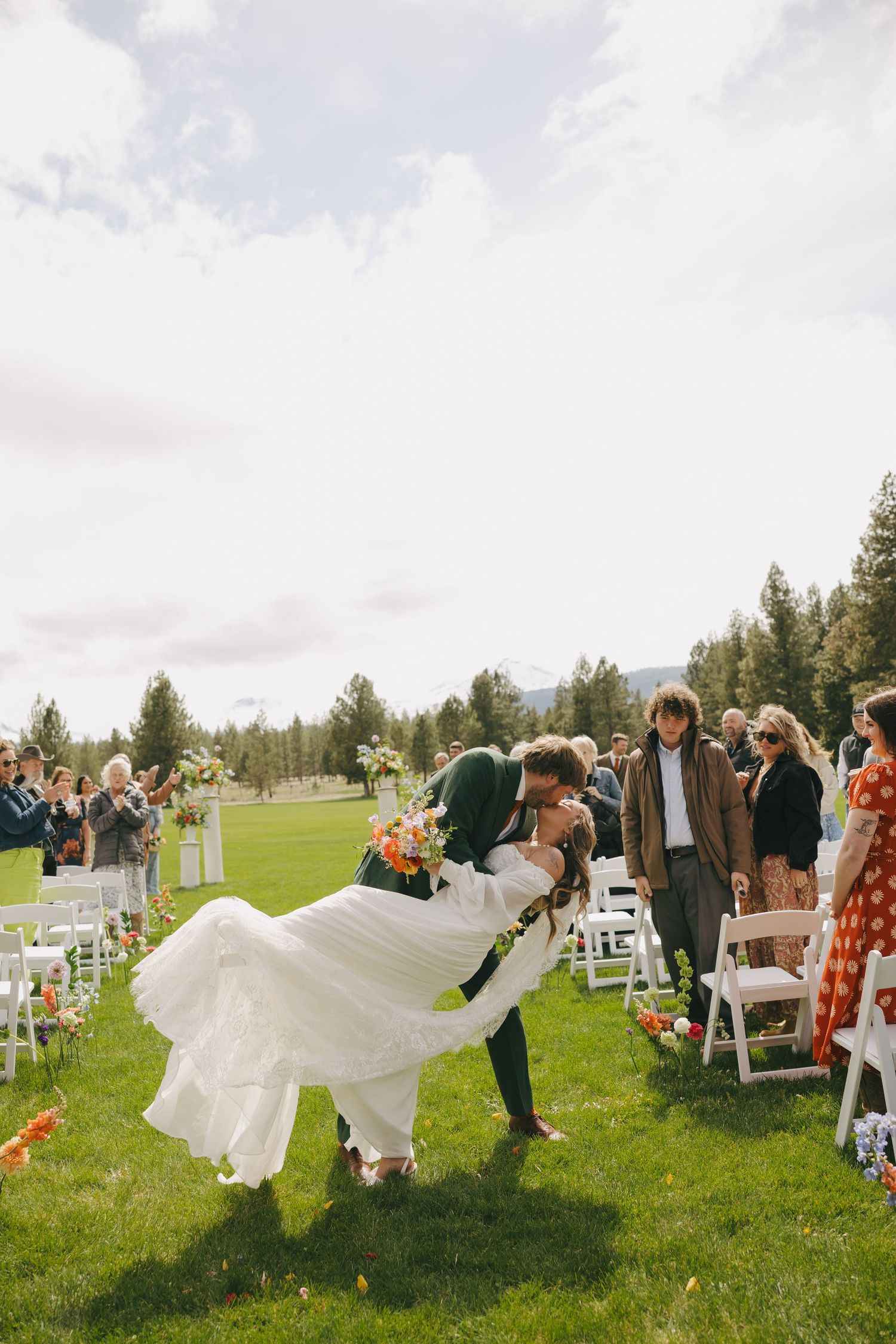 A couple celebrates at an outdoor wedding ceremony with guests seated on white chairs on a grassy lawn.