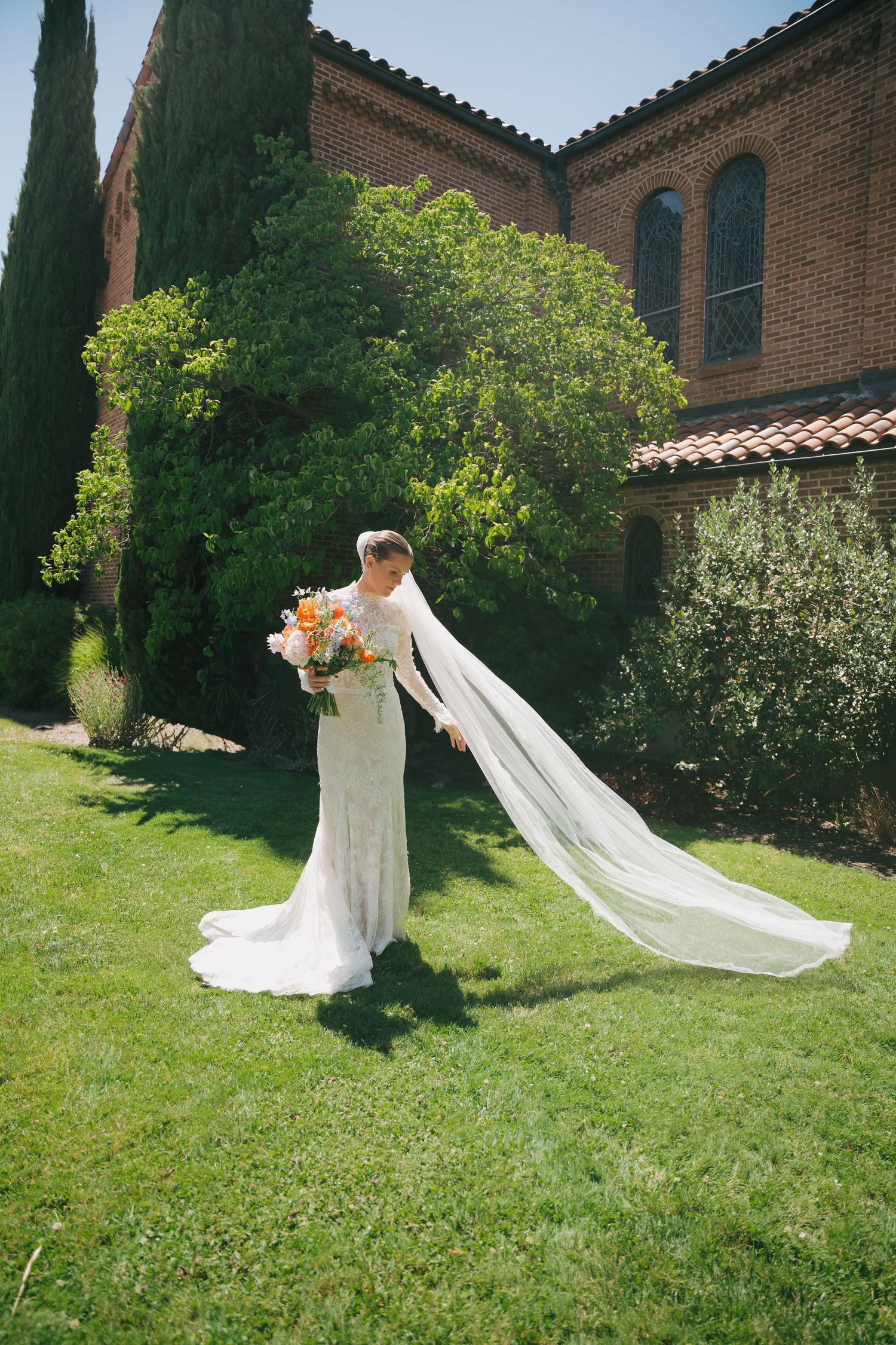 Bride in white gown with long veil holds colorful bouquet on green lawn beside brick church with cypress trees.