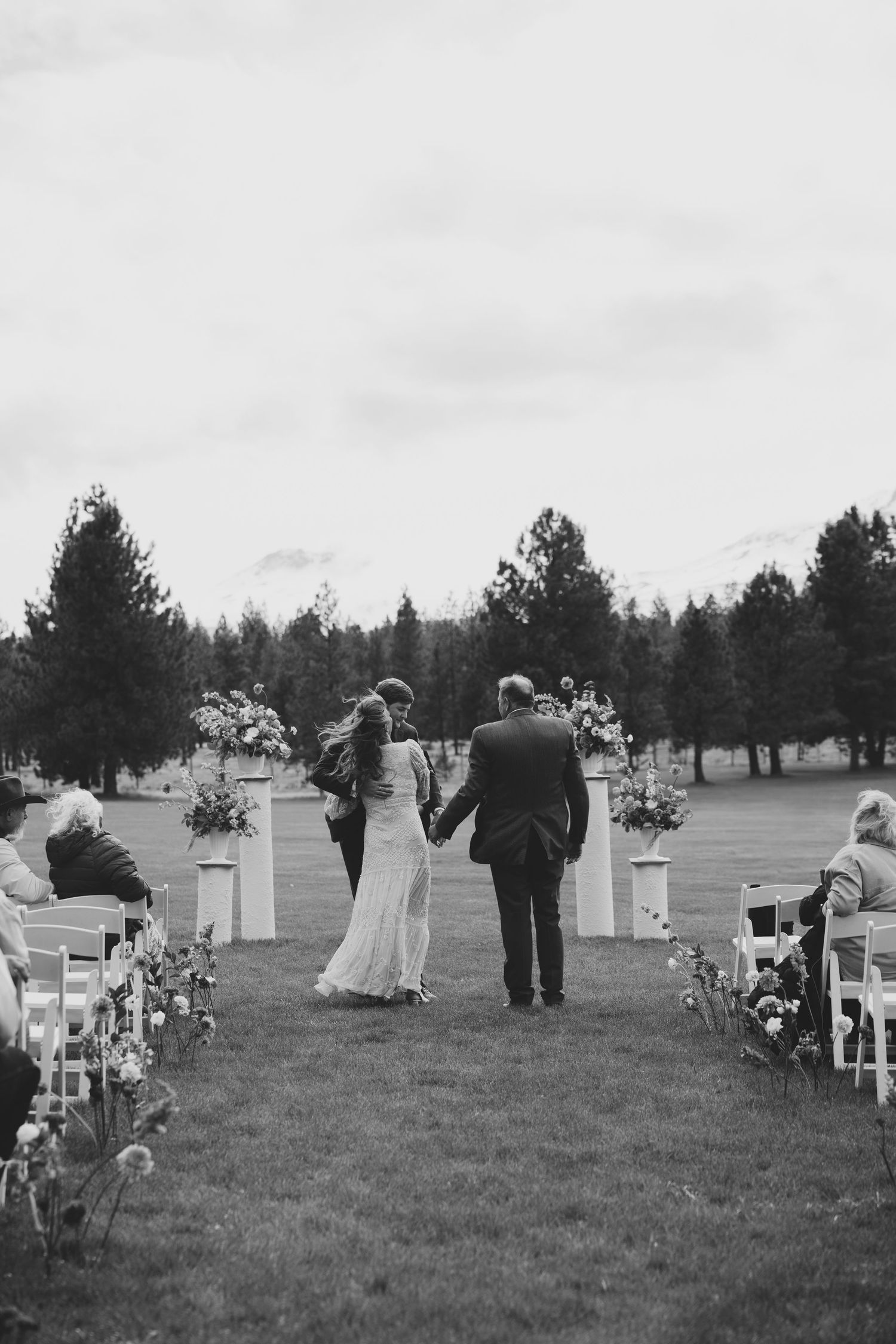 Black and white photo of an outdoor wedding ceremony setup with white chairs and floral arrangements on a grassy field.