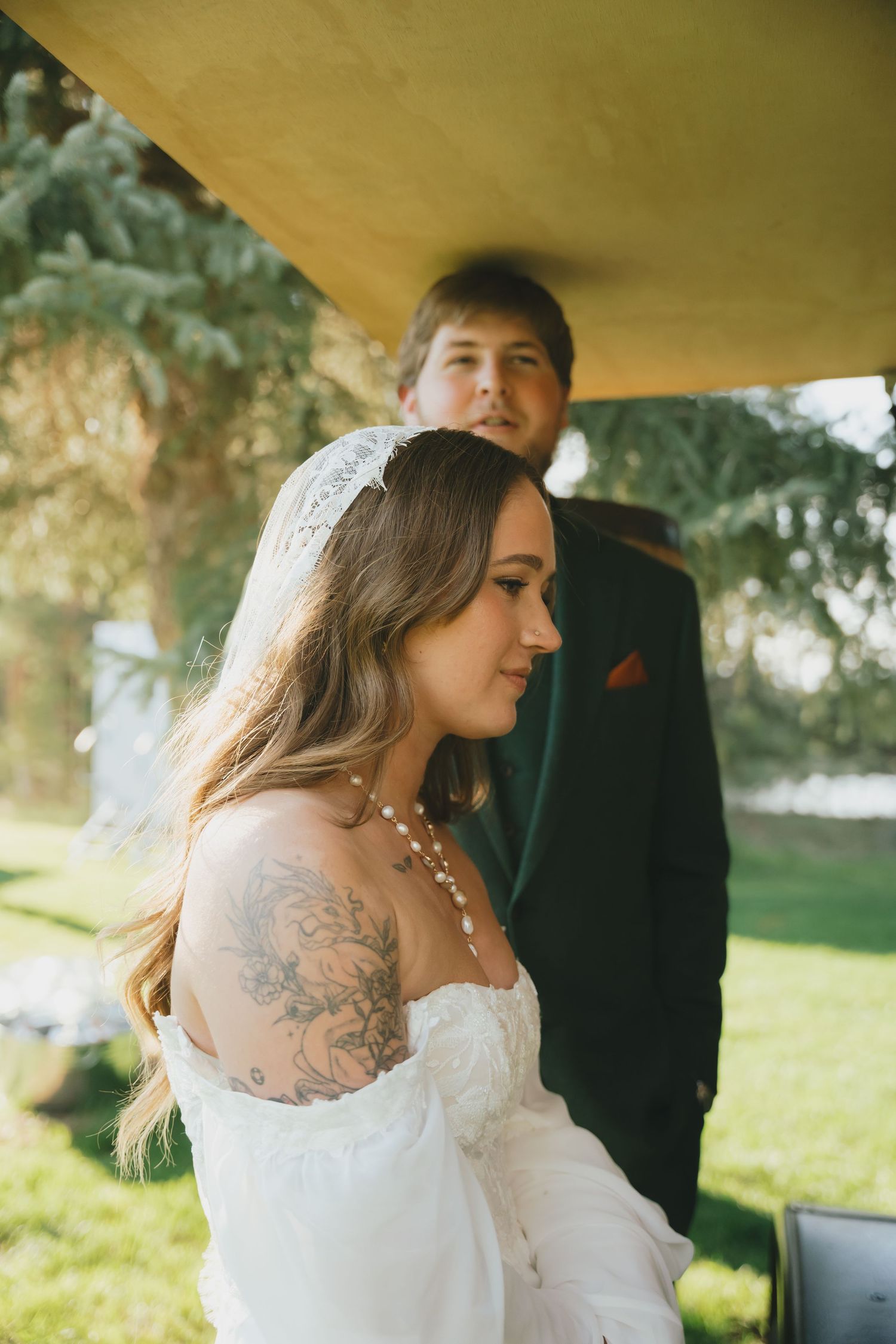 A bride in a lace wedding dress with tattoos stands in profile during an outdoor ceremony.