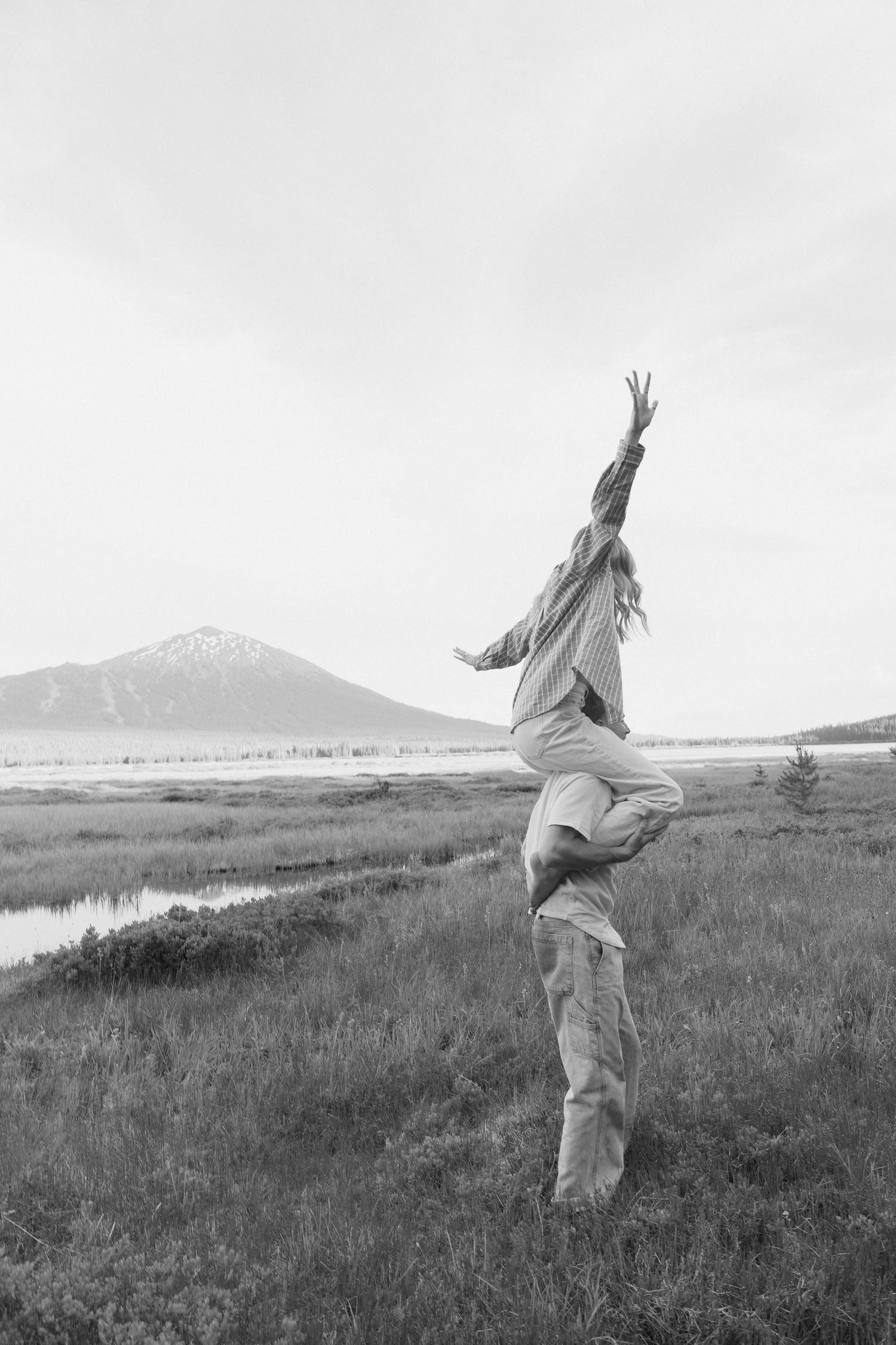 Black and white photo of a woman being lifted by a man in a scenic mountain meadow with a river in the background.