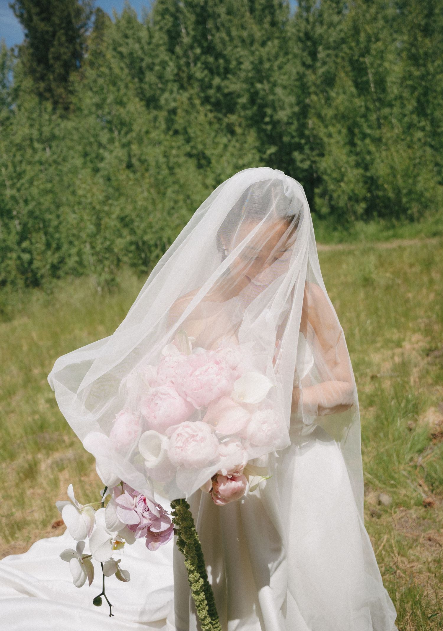 Bride in white gown and veil holding a lush pink peony bouquet outdoors with green trees in background.