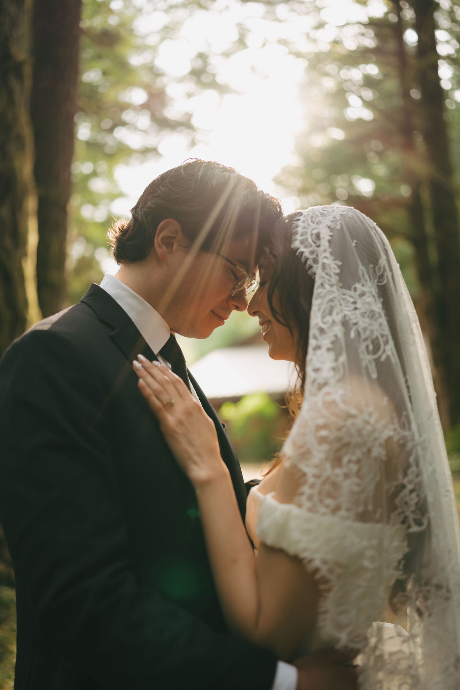 Bride and groom touching foreheads in a sunlit forest, bride wearing lace veil and gown, groom in black suit.