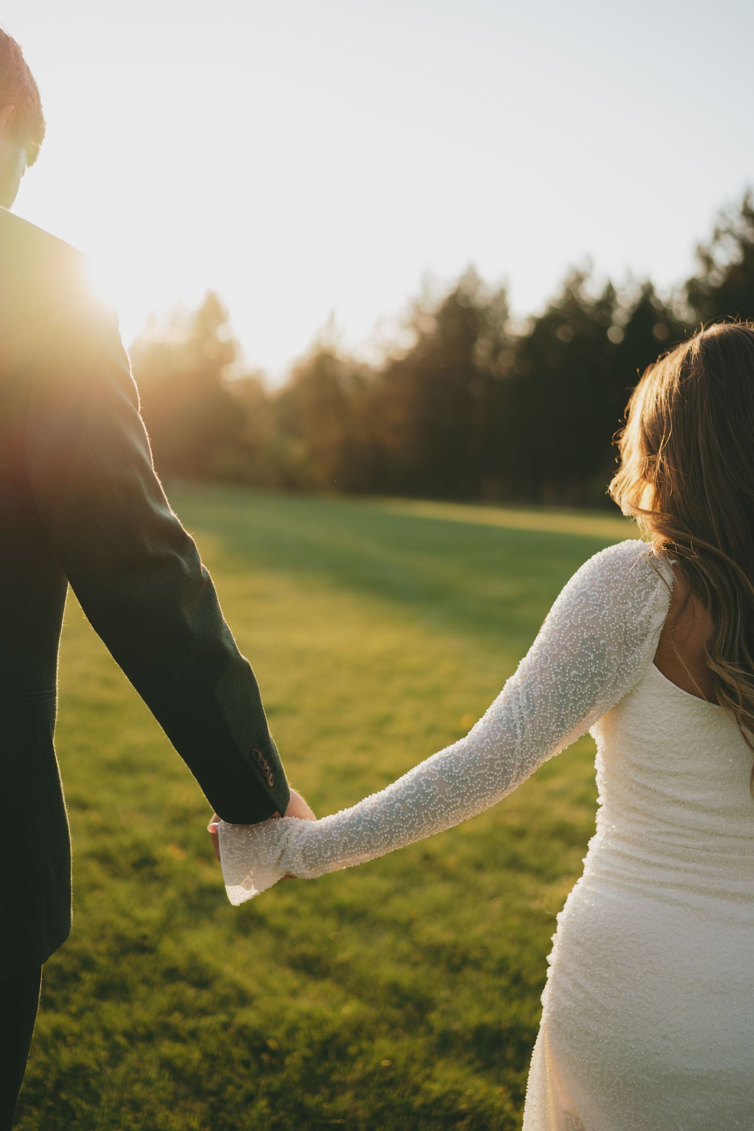 A couple holds hands in a grassy field during sunset, with the woman wearing a white dress showing her pregnancy.