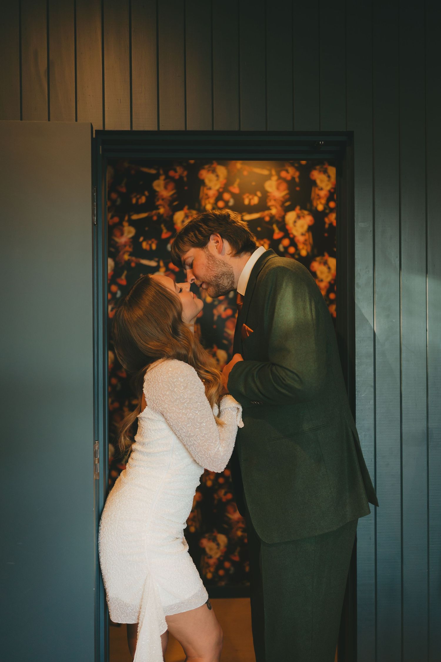 A couple in formal attire shares a romantic moment against a dark wall with glowing orange lights in the background.