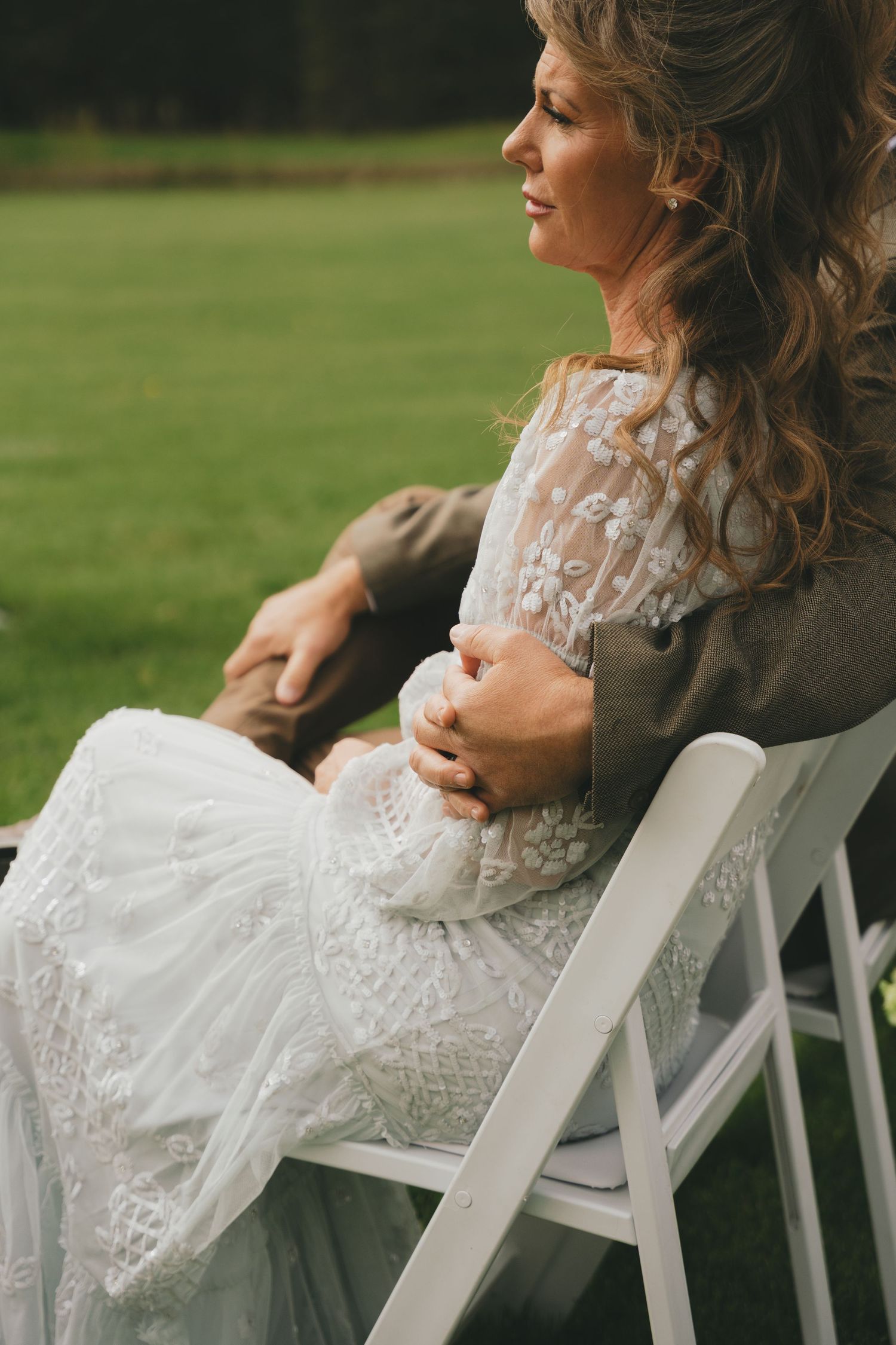 A person in a white lace dress sits on a white folding chair outdoors with a baby in their arms.