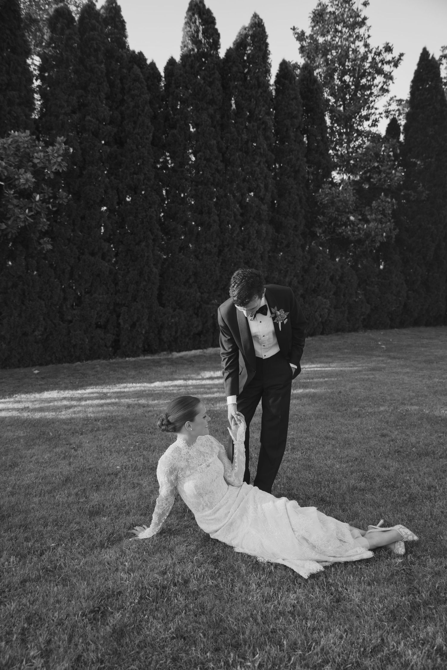 Black and white photo of a bride in lace gown sitting on grass while groom in tuxedo stands beside her outdoors.