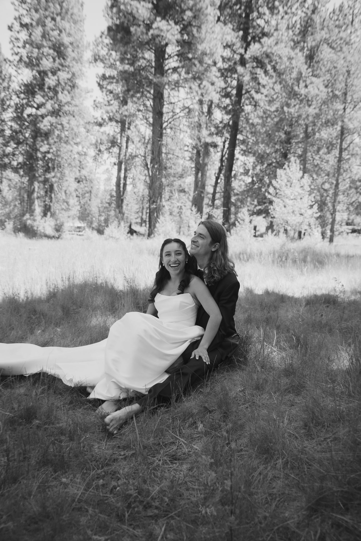 Black and white photo of a laughing bride in white gown sitting with partner on grass among tall pine trees.