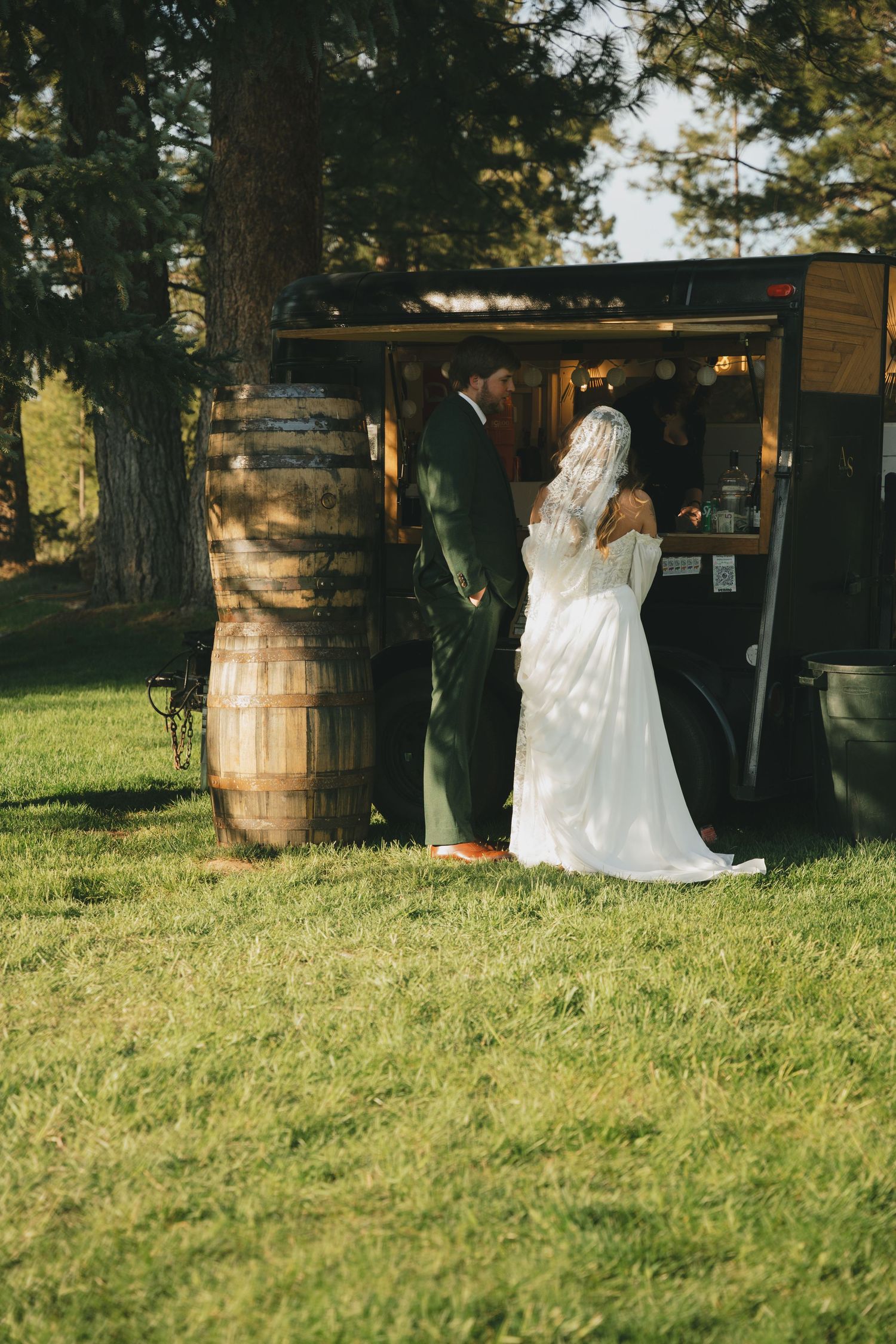 A couple in wedding attire stands near a rustic wooden barrel outdoors at a countryside venue.