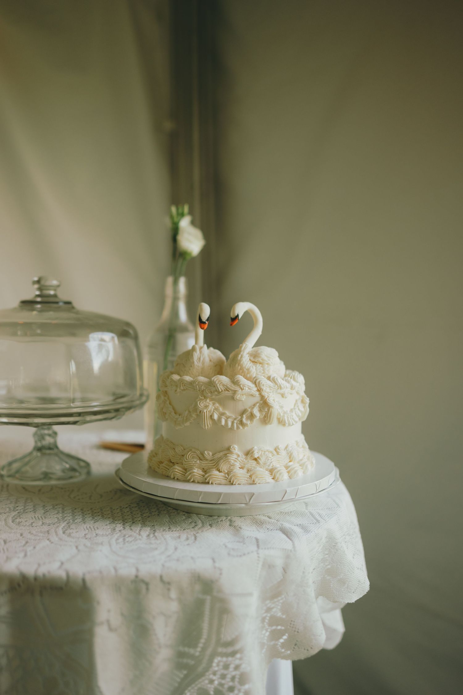 Small white wedding cake with lace details and swan topper on lace-covered table with glass dome.