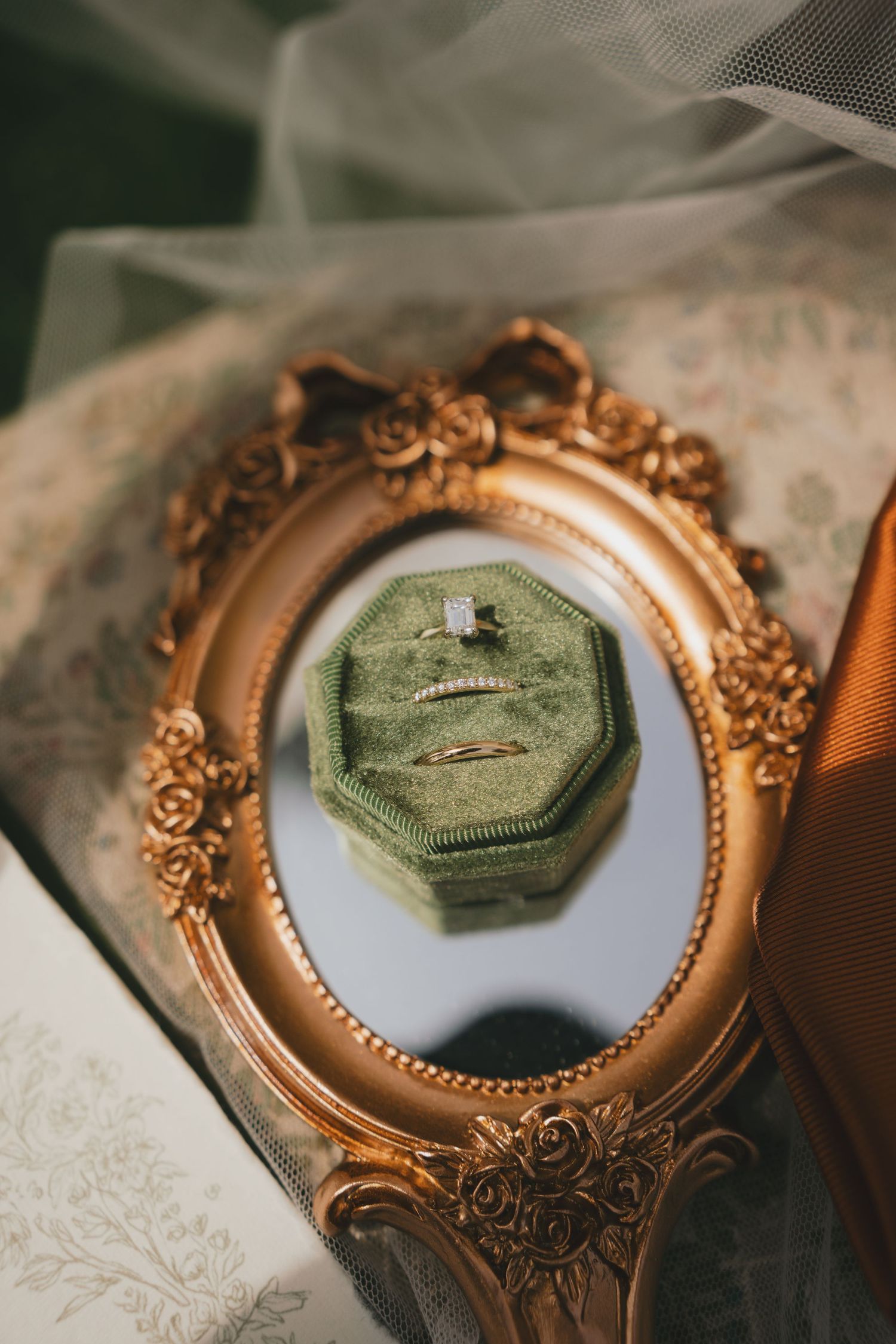 Ornate gold vintage mirror frame displays a green velvet ring box on white lace fabric.
