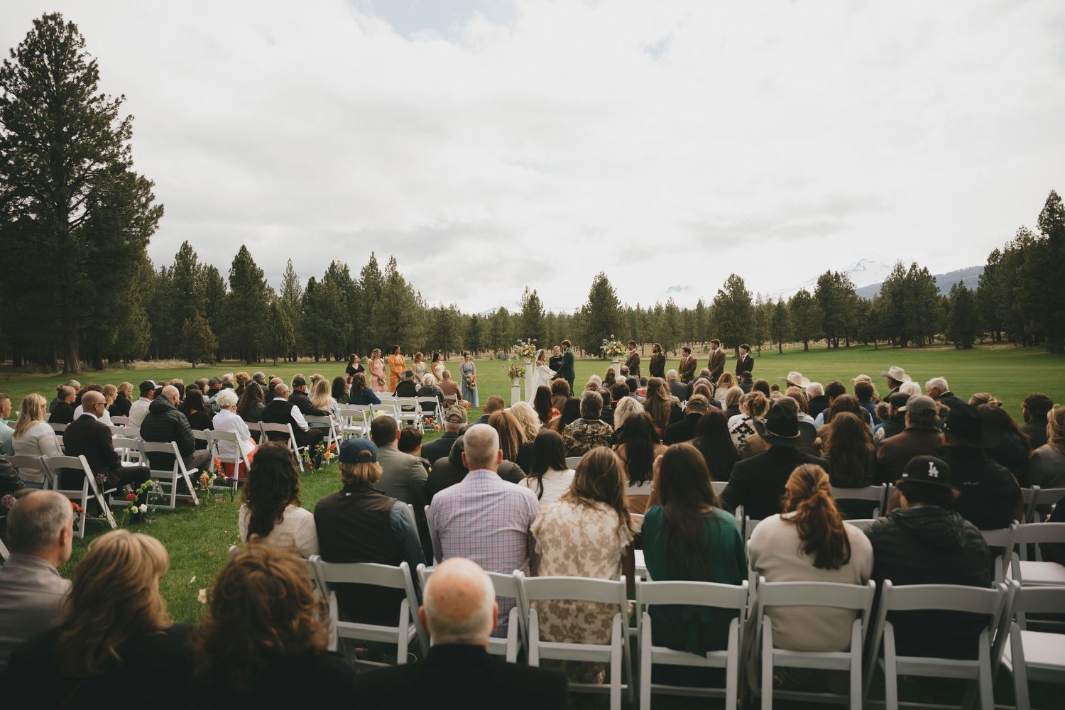 Panoramic view of outdoor wedding ceremony with white chairs set up on green lawn surrounded by evergreen trees.