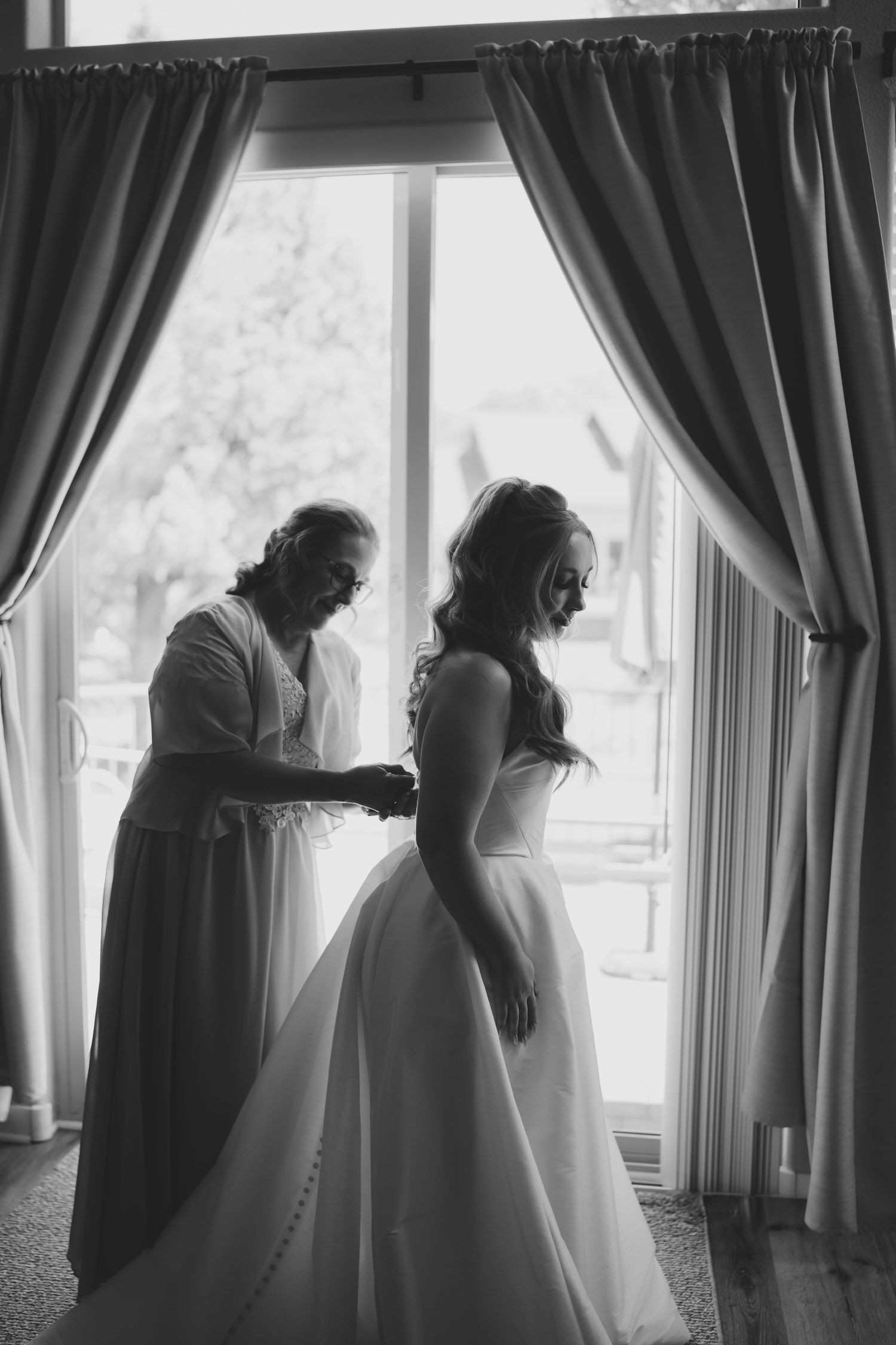 Black and white photo capturing wedding dress preparation by window with elegant curtains.