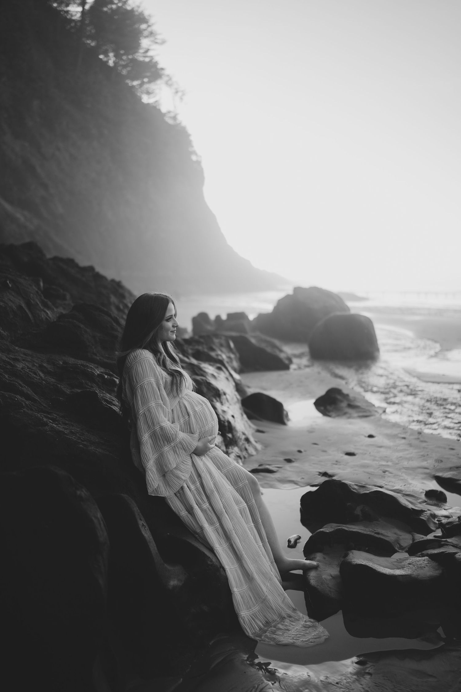 A dramatic black and white photo series of a person in a flowing white dress on a rocky beach at sunset.