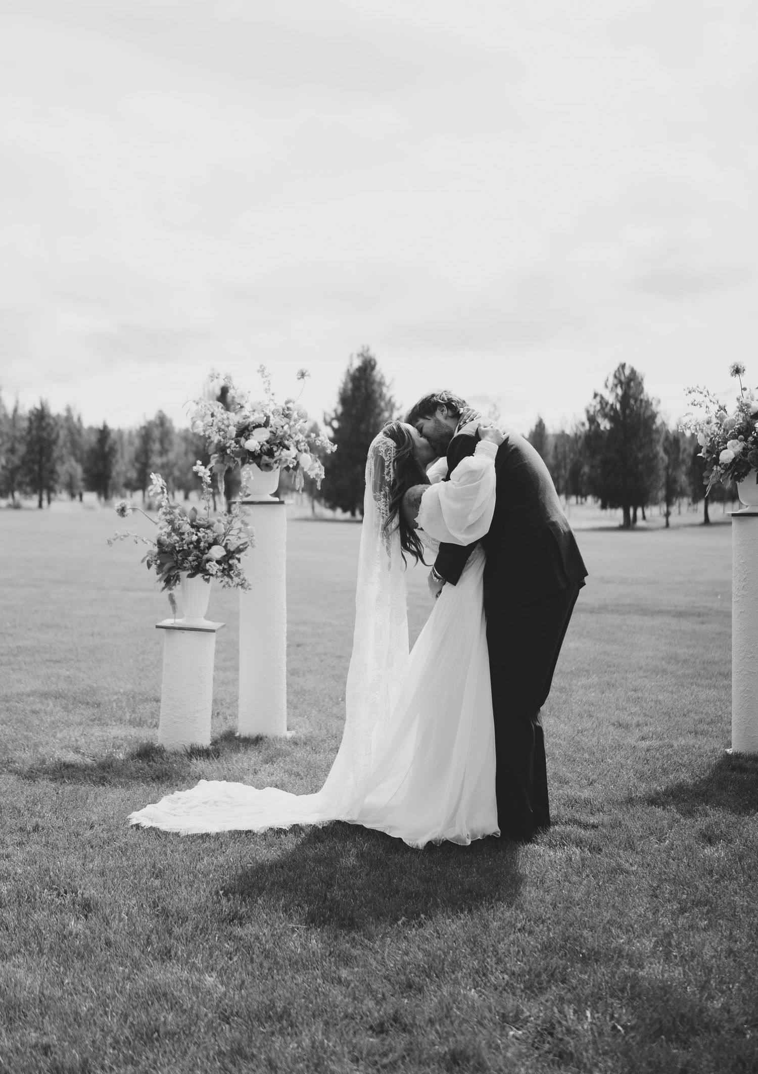 Black and white wedding photo showing couples lined up for a ceremony in an open field with decorative pillars.