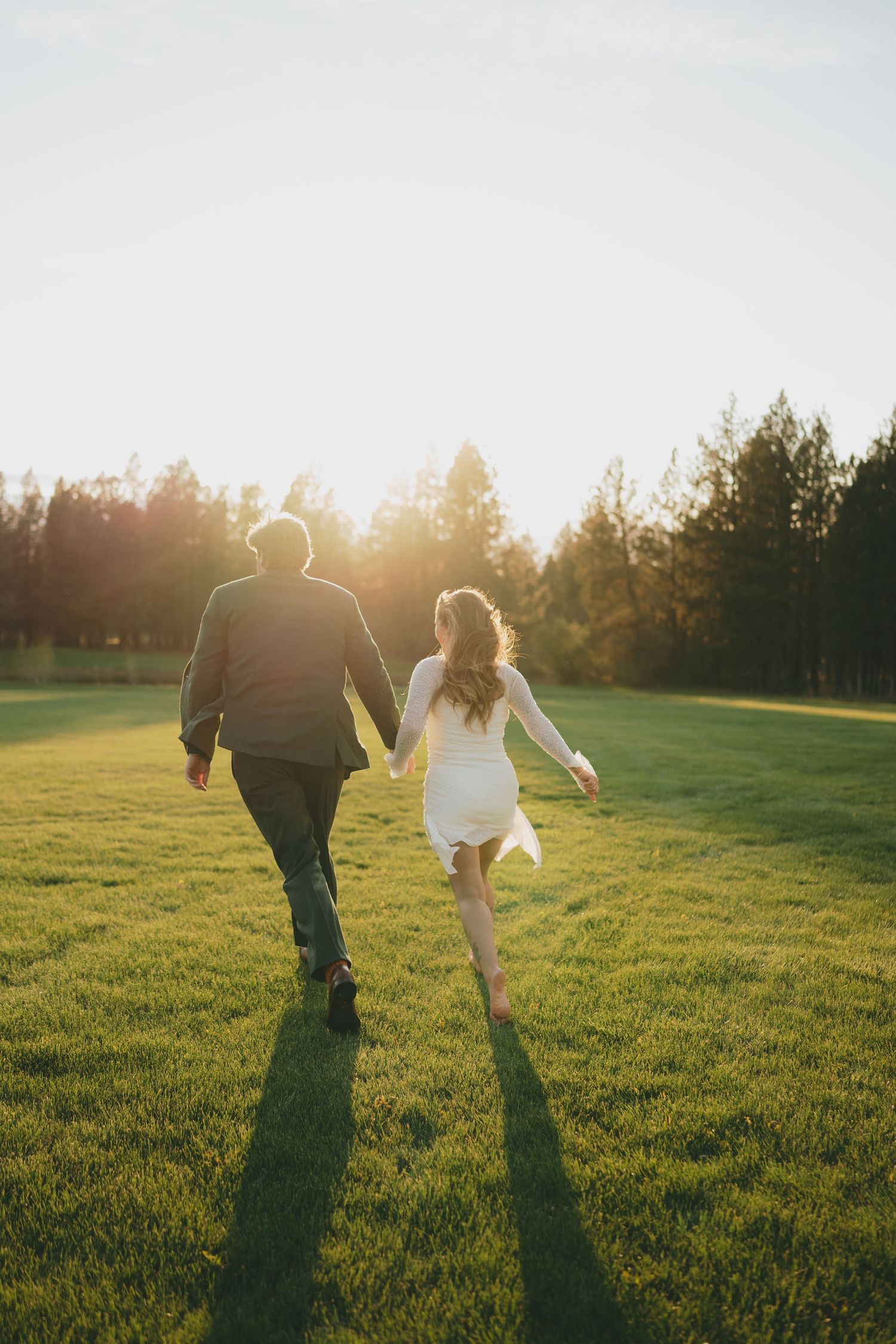 A couple runs playfully through a sunlit meadow at golden hour, casting long shadows across the green grass.