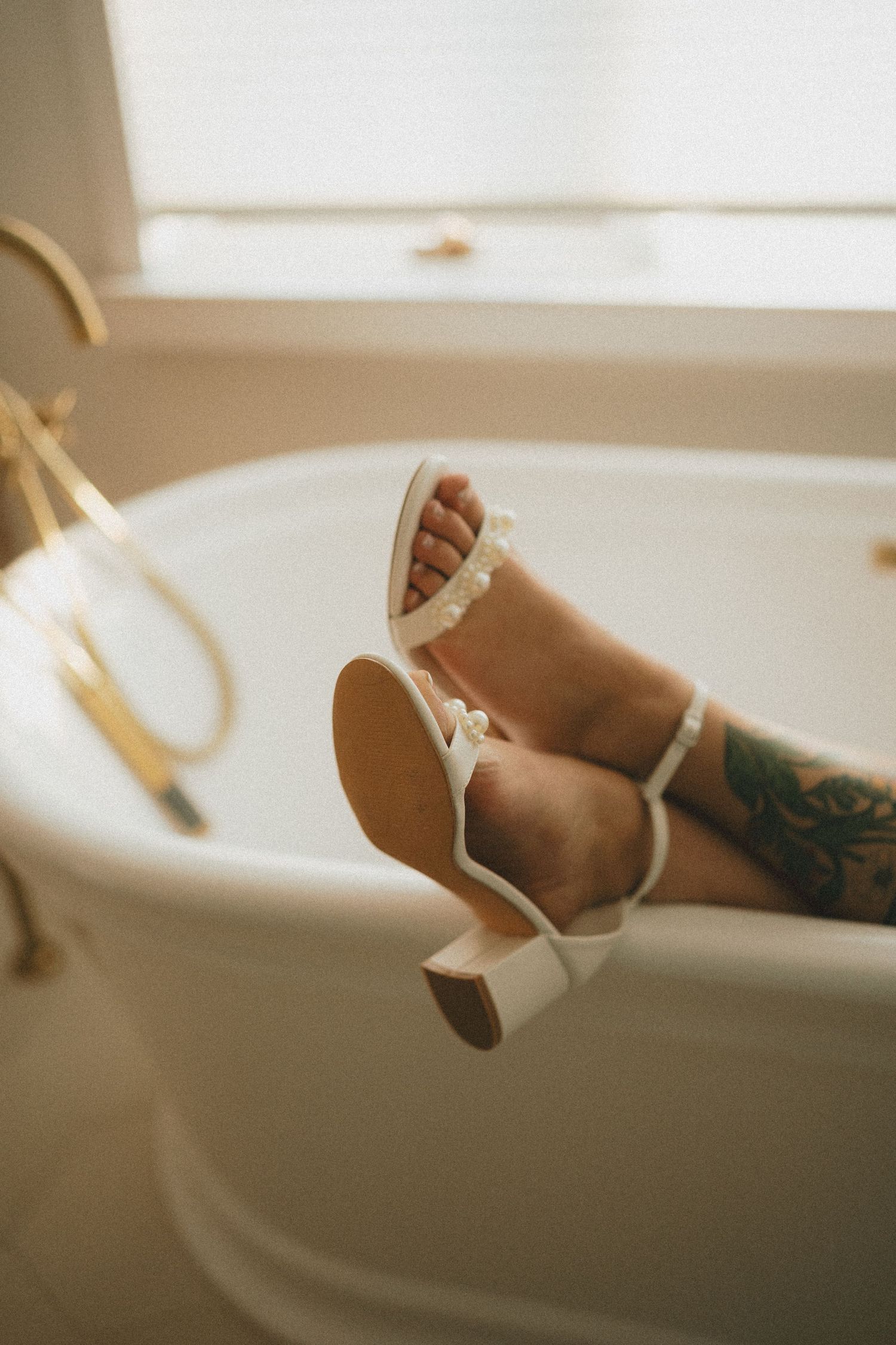 Feet wearing delicate white sandals peek over the edge of a freestanding bathtub.