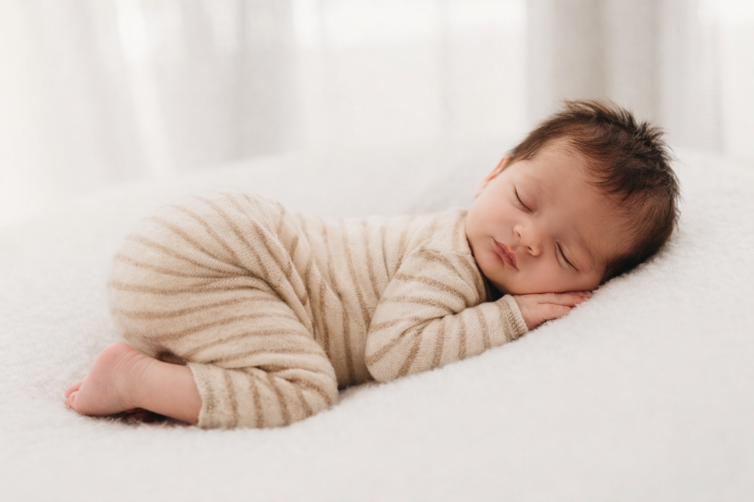 Sleeping newborn in beige outfit curled up on white blanket.
