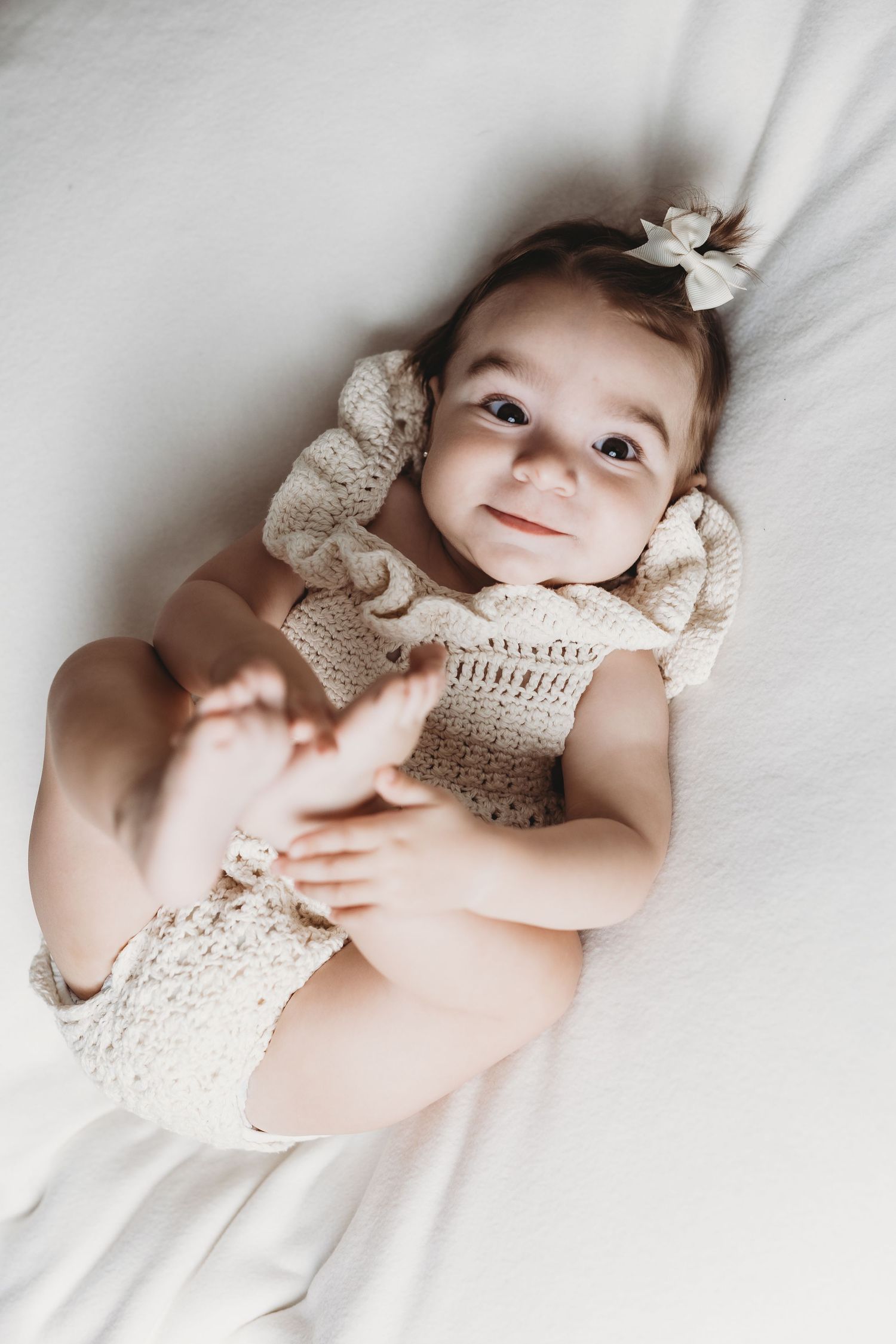 Baby wearing white lace dress lies on white bedding while smiling.