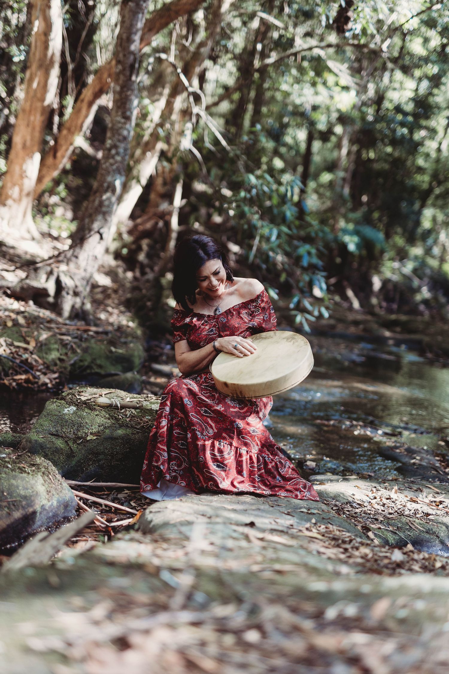 Person in red dress playing drum in forest creek setting.