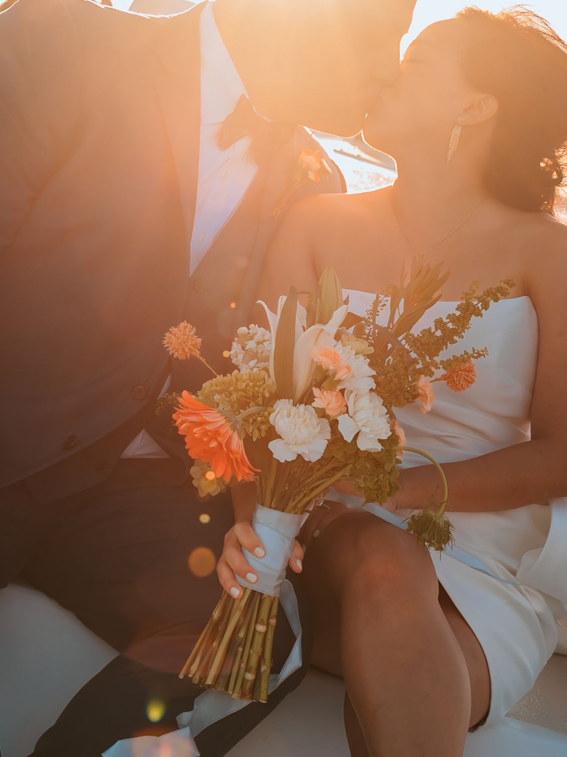 Couple kissing at golden hour wedding holding a vibrant orange and white bridal bouquet in warm sunlight.