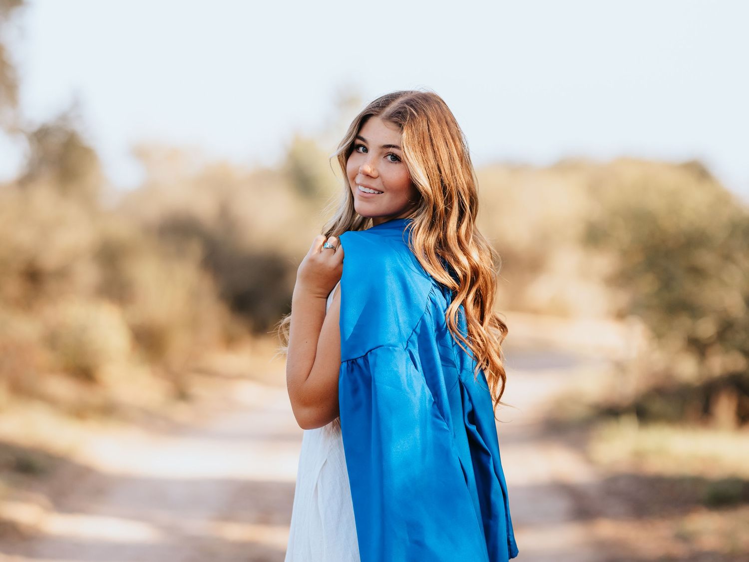 Young woman with long wavy hair in a blue dress smiling over her shoulder on a sunny dirt path.