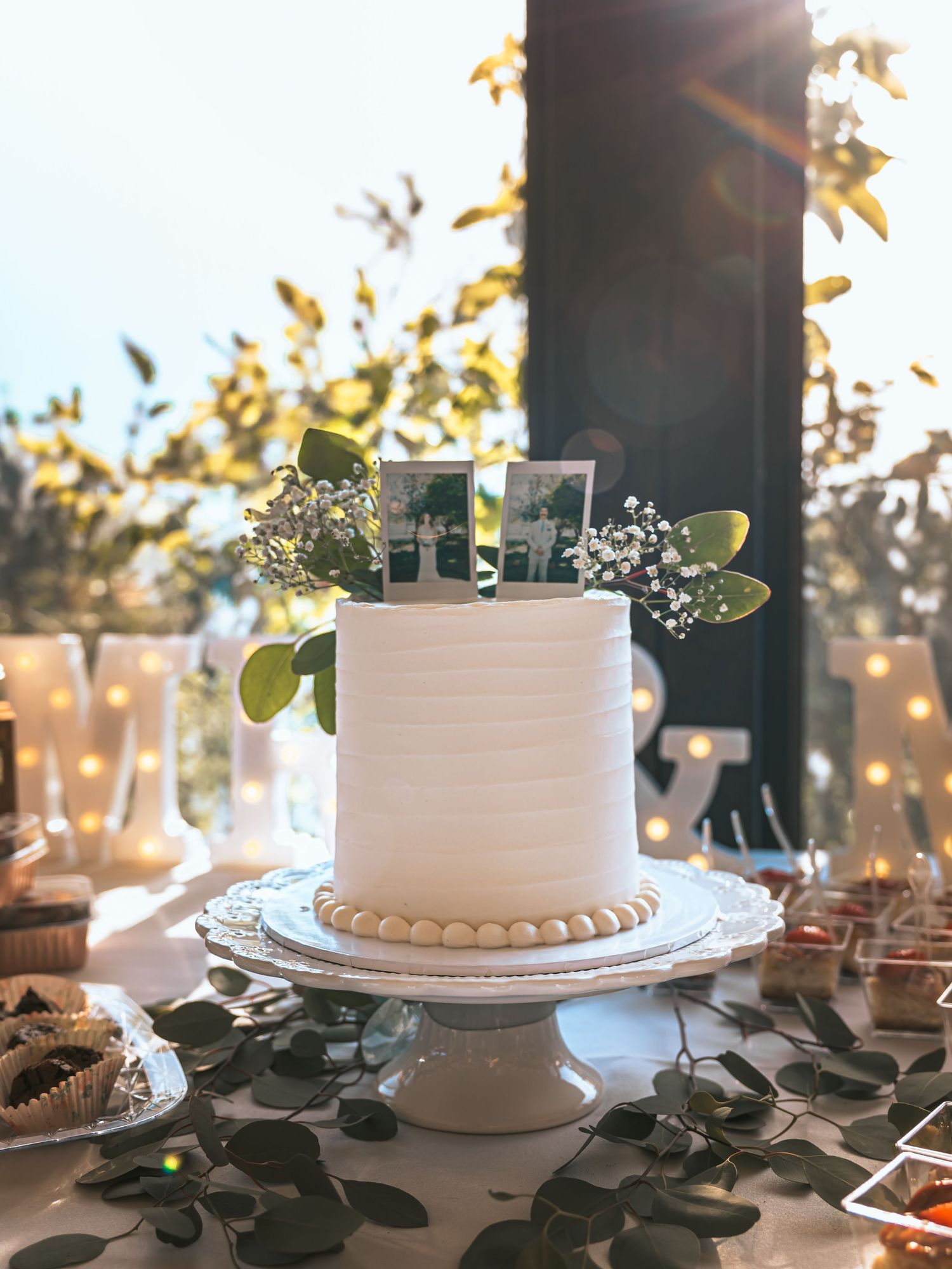 Elegant single-tier white wedding cake with floral topper on decorated dessert table with greenery.