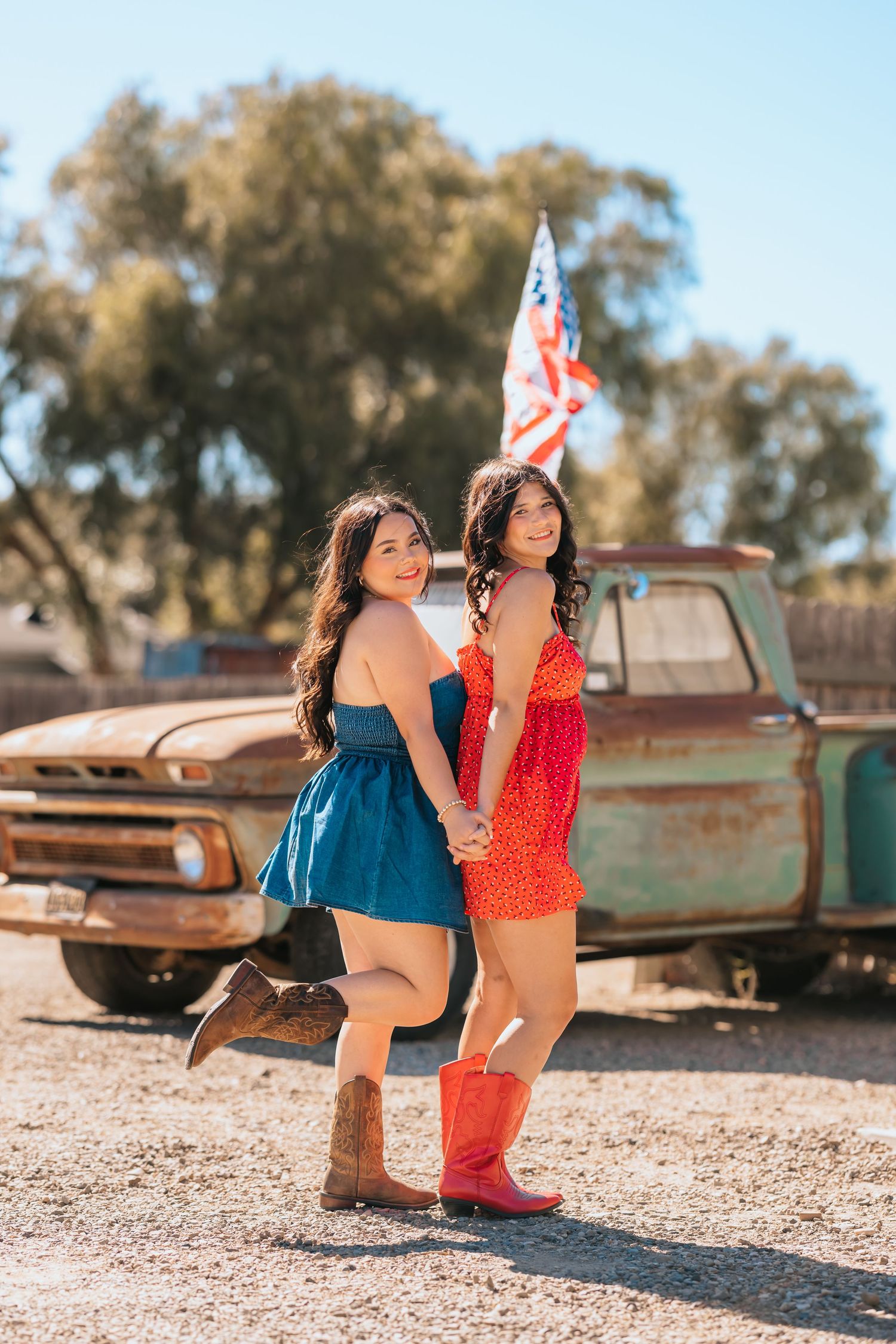 Two young women in colorful dresses and cowboy boots pose in front of a rusty vintage truck.