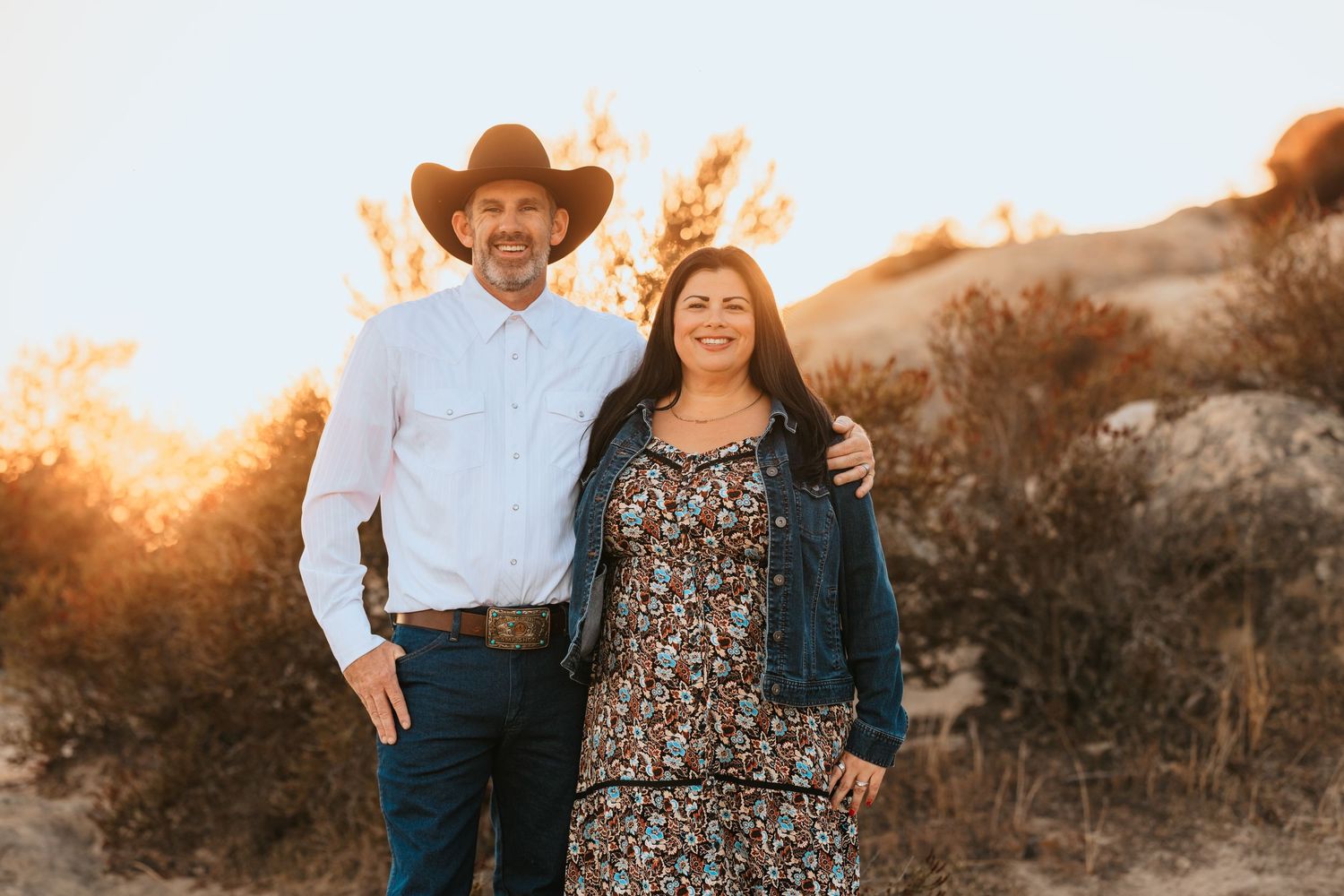 Couple posing outdoors at golden hour, man in cowboy hat and woman in floral dress with denim jacket.