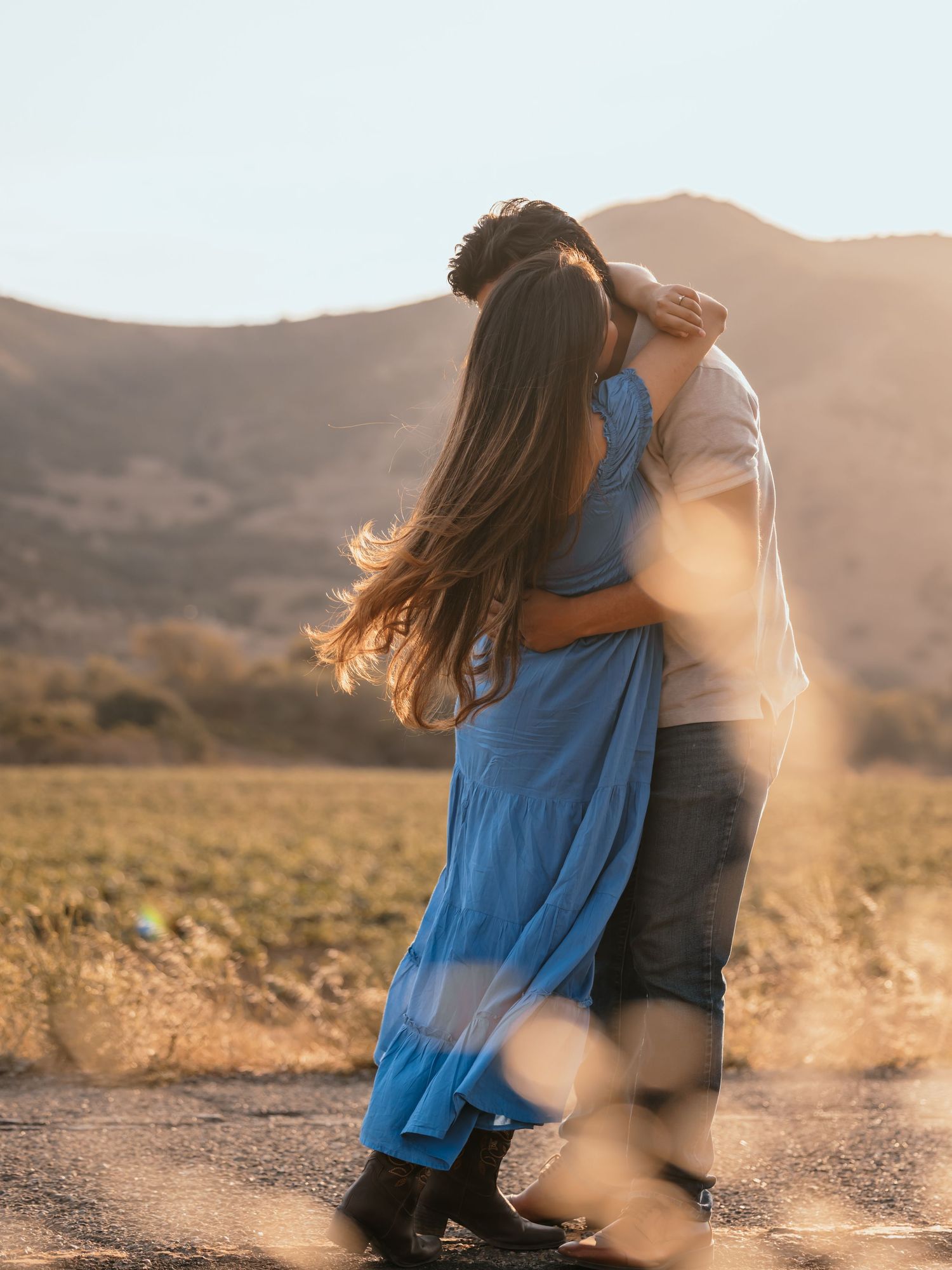 A couple embraces and kisses at golden hour on a dirt road with rolling hills and warm sunlight behind them.