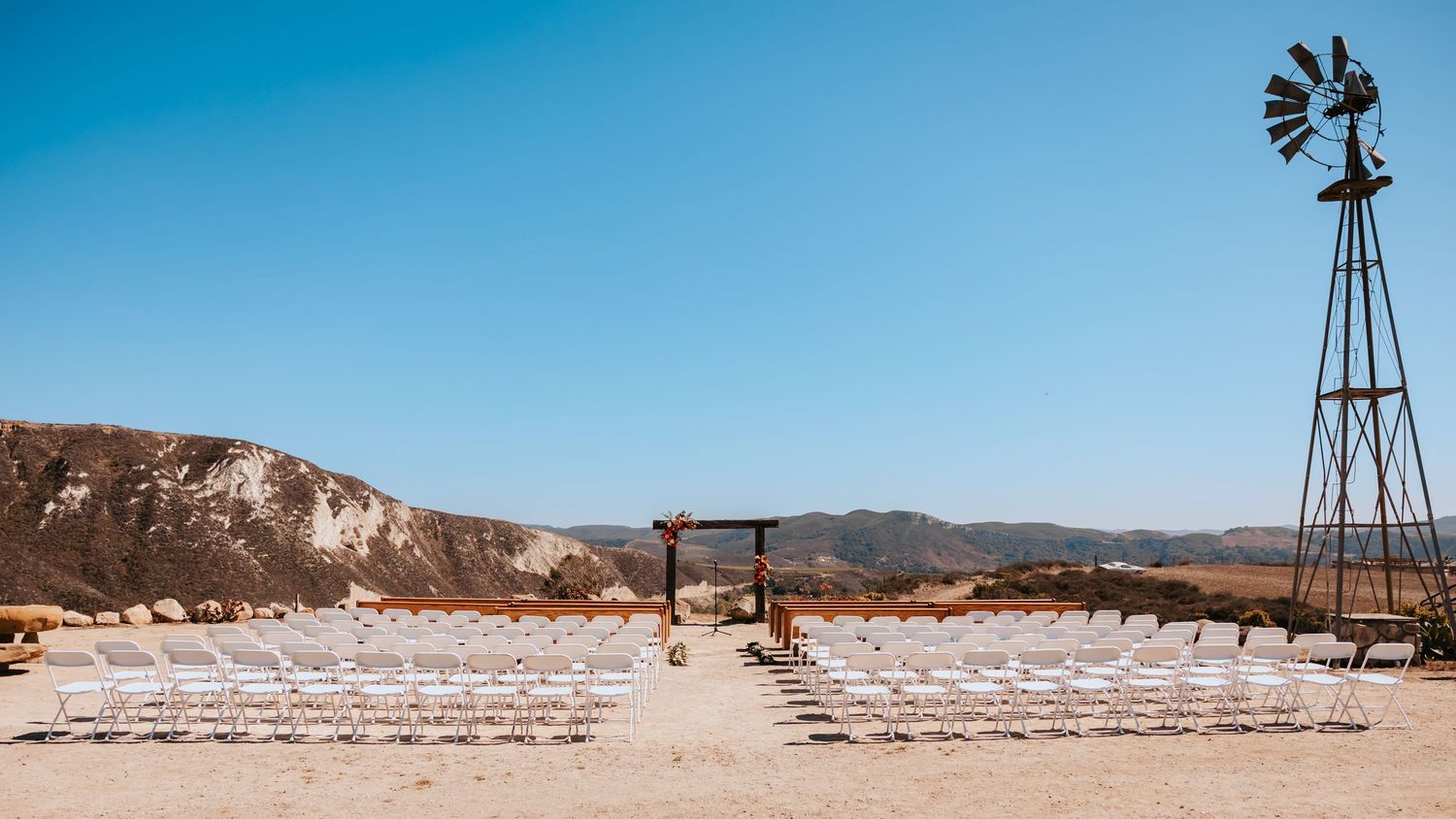 A panoramic desert landscape with windmills and white chairs arranged for an outdoor event against an arid backdrop.