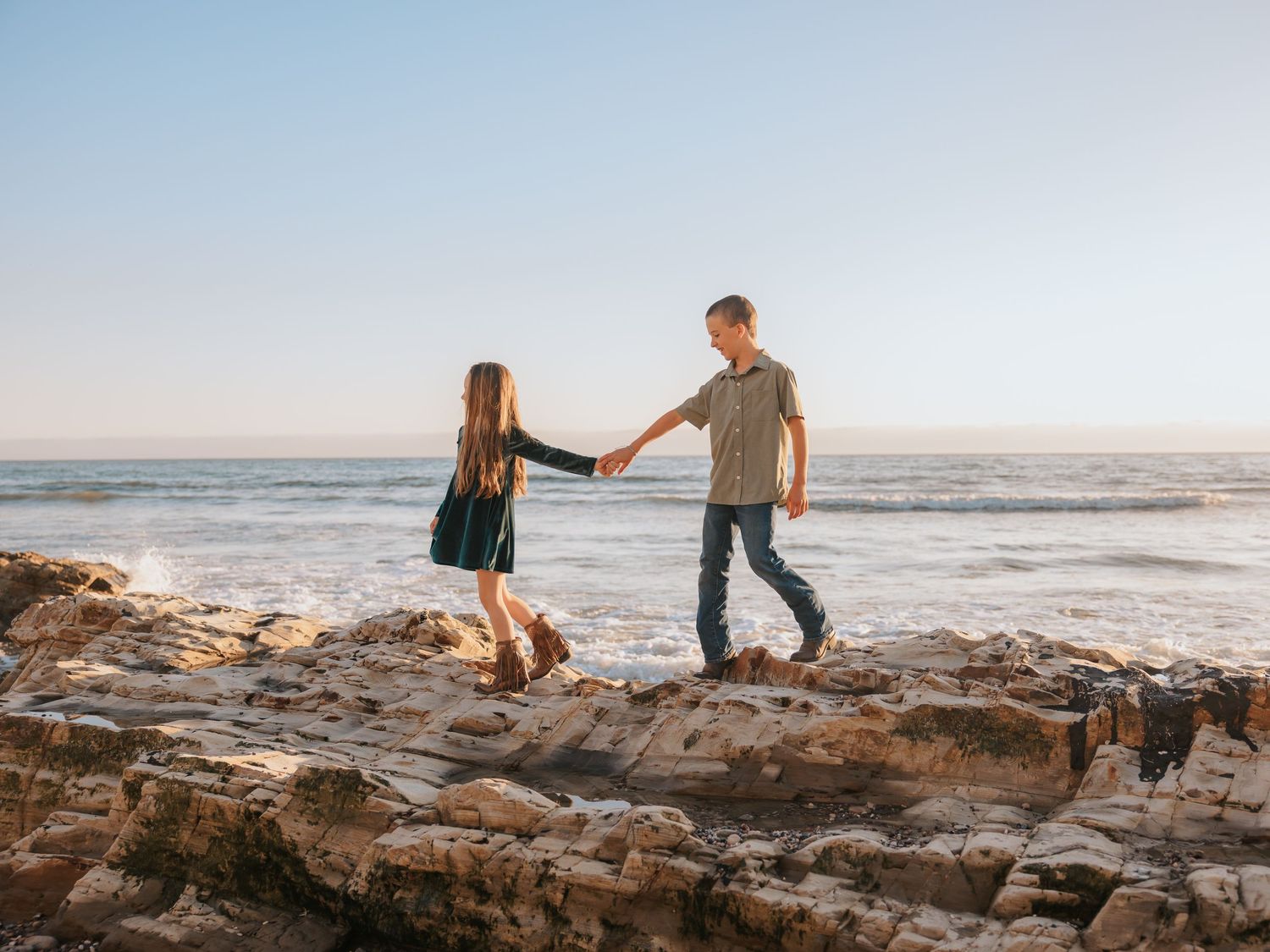 Two people holding hands while walking together on rocky coastal shore at sunset by the ocean.