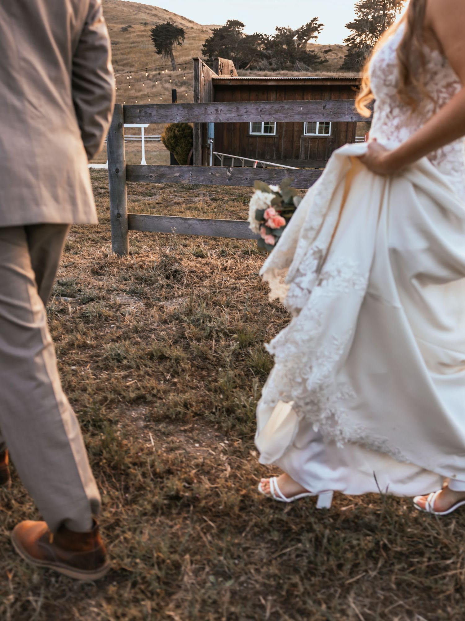 Bride lifting lace gown hem while walking with groom near rustic barn fence at golden hour.