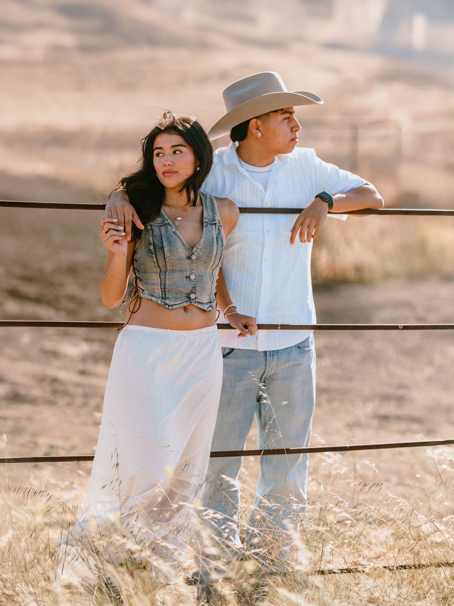 A couple in western attire leans on a fence in a dry field, woman in crop top and white skirt, man in cowboy hat.