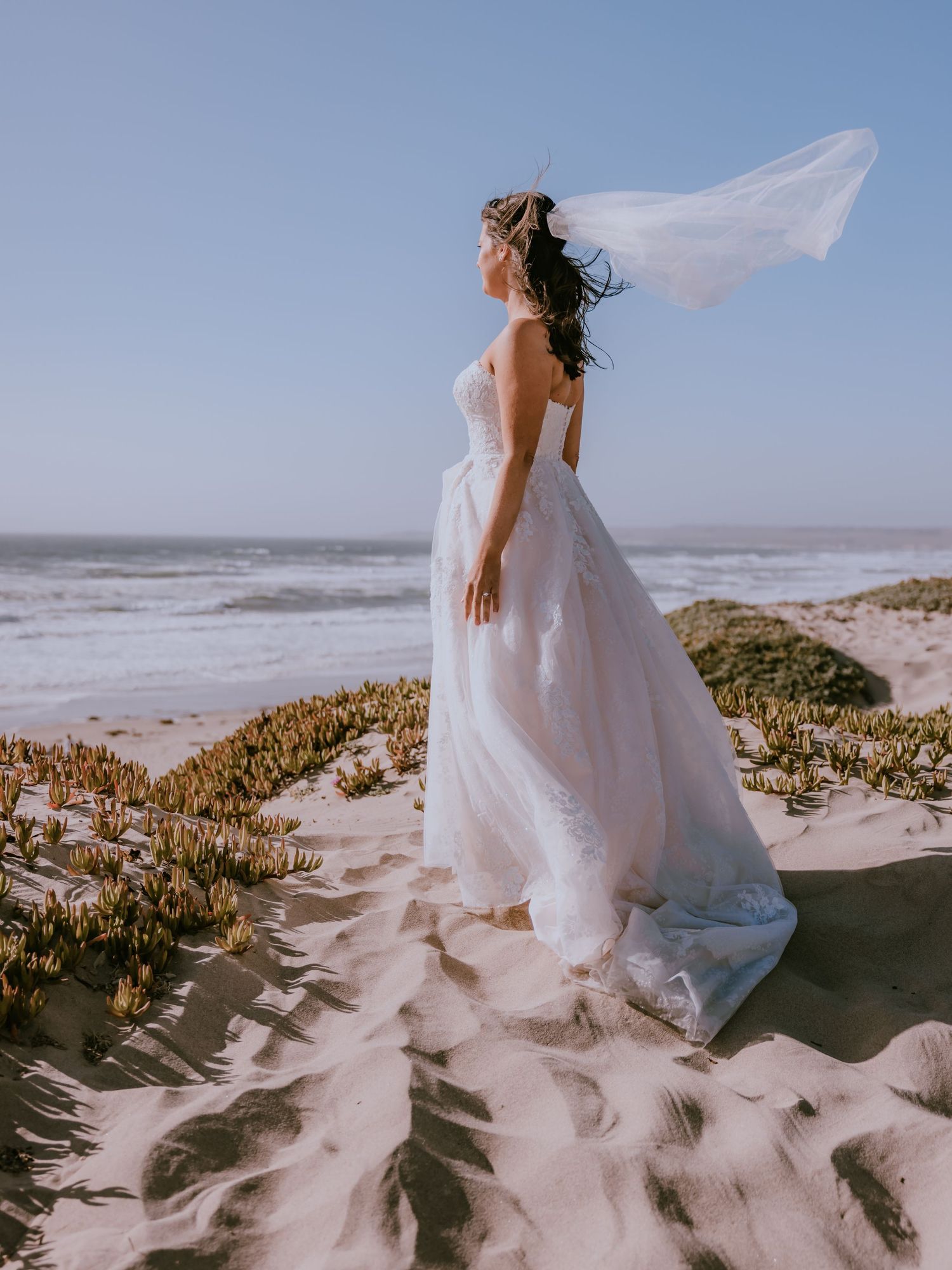 Ethereal white wedding dress flows in ocean breeze as figure stands gracefully on sandy beach dunes at sunset.