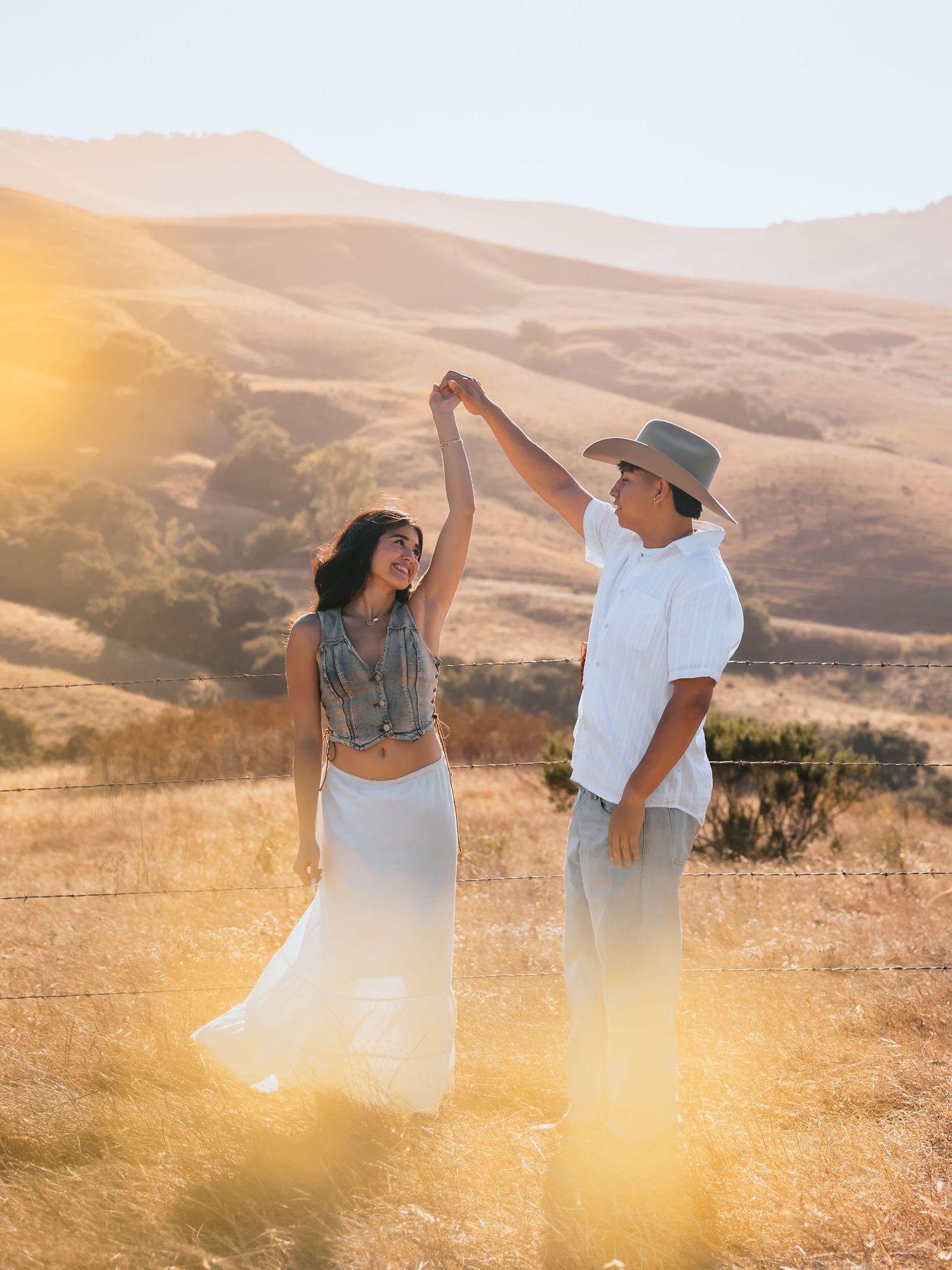 Couple dancing at golden hour in a sunlit field with rolling hills, woman in white skirt, man in hat and white shirt.