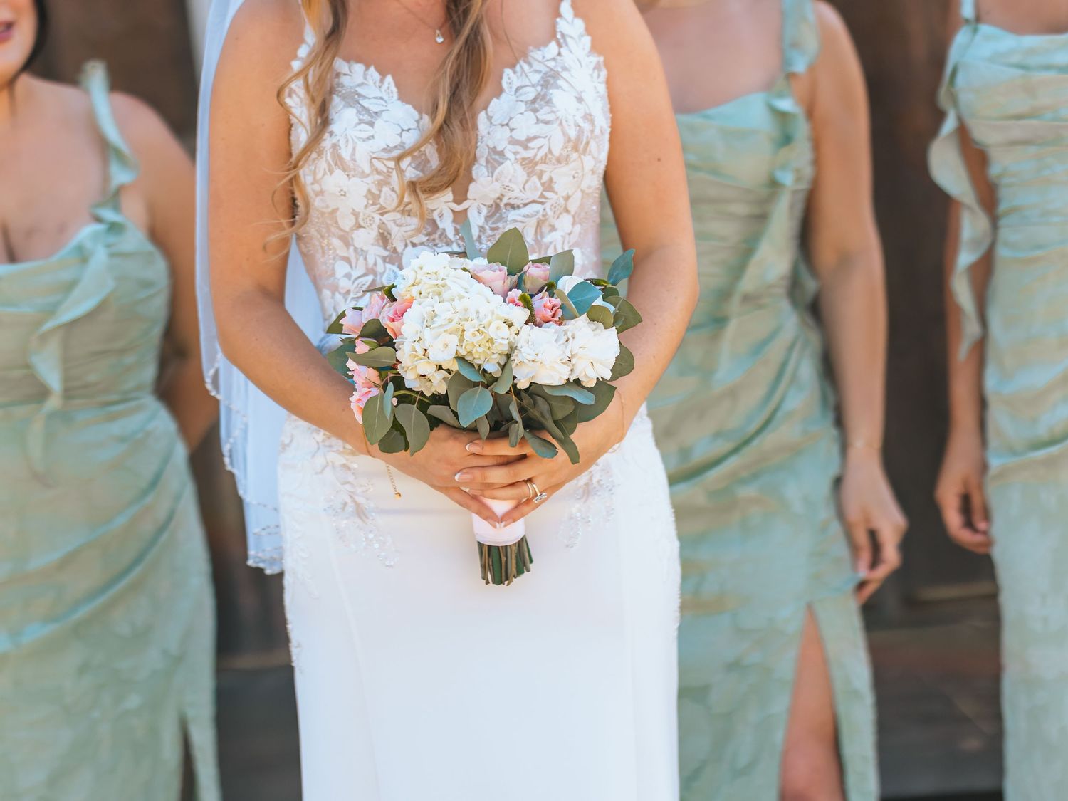 Close-up of bride in lace gown holding white and pink floral bouquet with bridesmaids in sage.