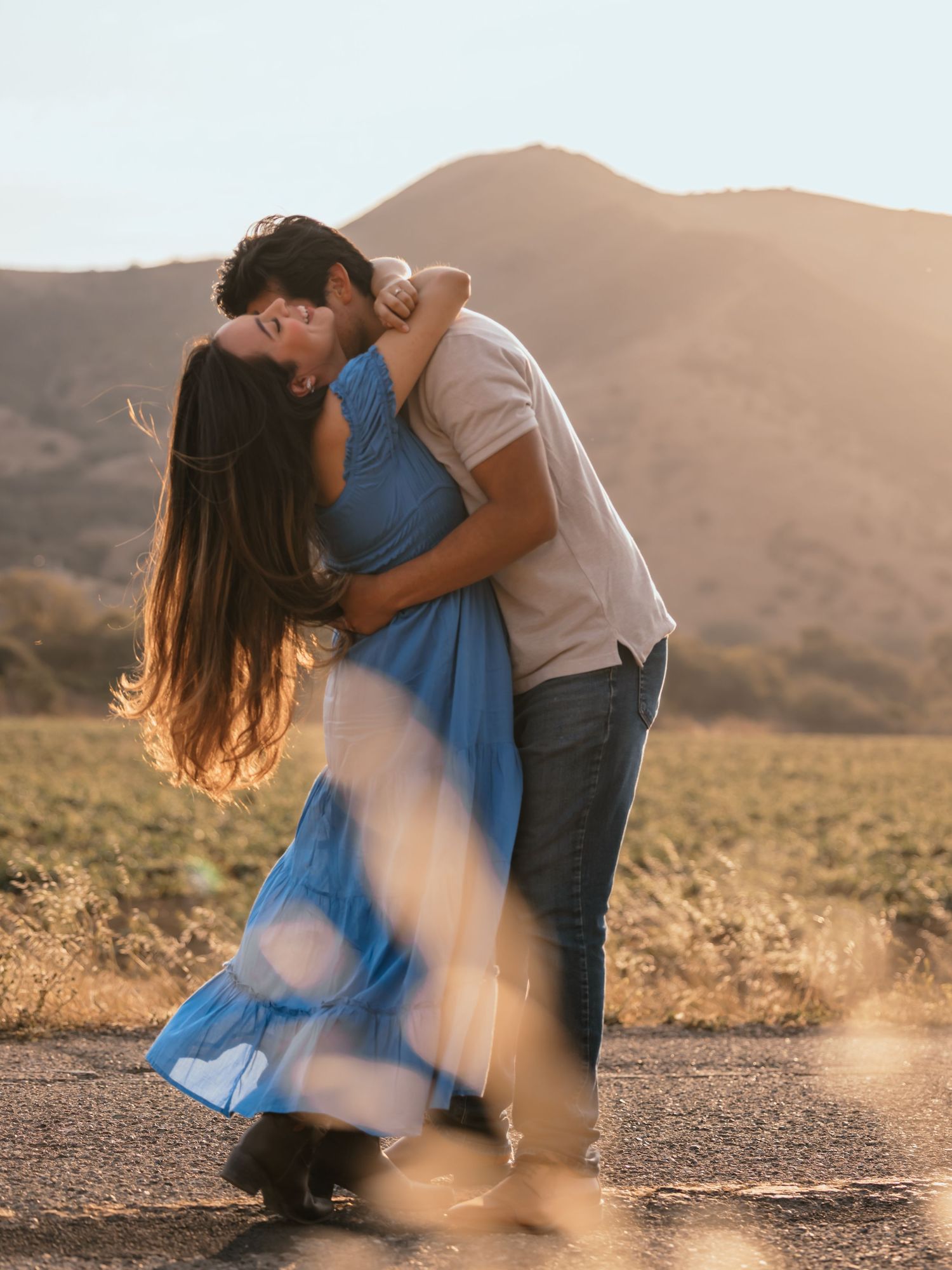 Couple sharing a romantic kiss at golden hour on a road with rolling hills and warm sunlight in the background.