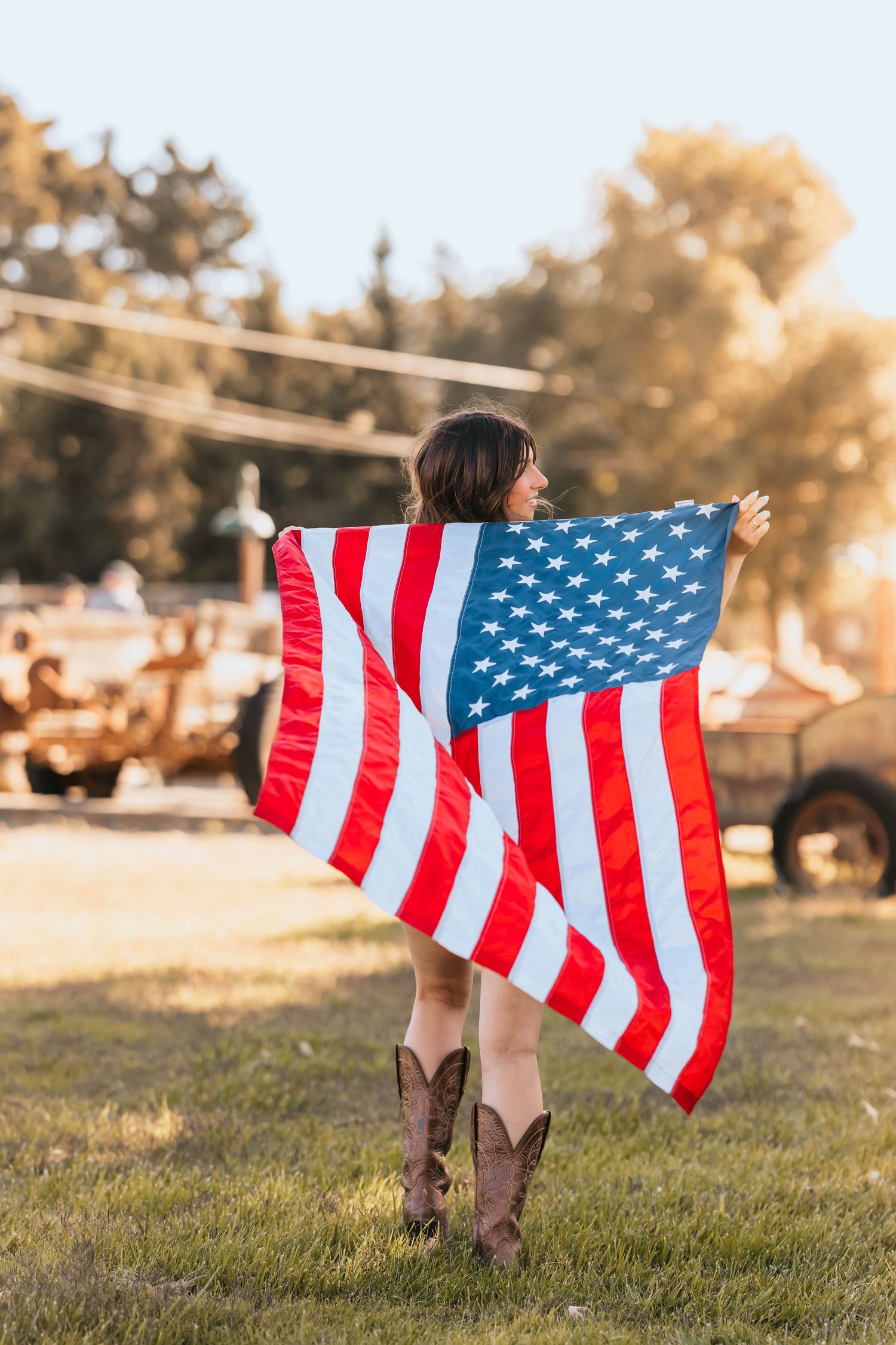 Woman holding large American flag wearing cowboy boots standing in sunny outdoor field.