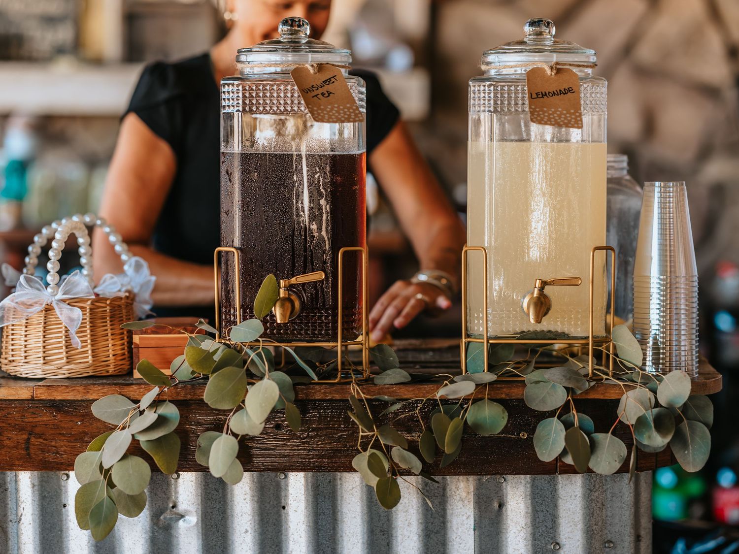 Two glass beverage dispensers with spigots decorated with eucalyptus leaves on a rustic wooden table at a drink station.